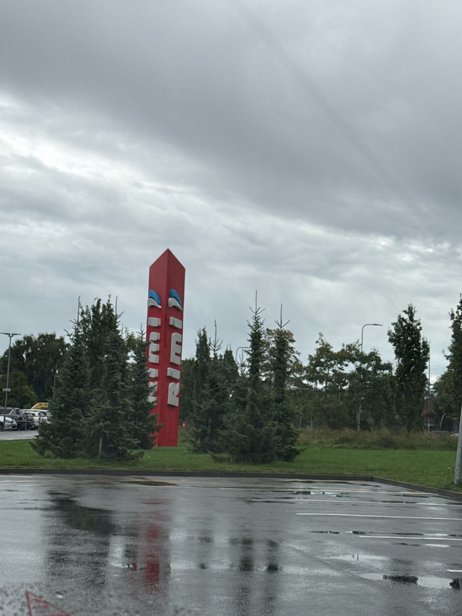 The image shows a large, red Rimi supermarket sign with white lettering and blue accents, positioned vertically. The sign is located outdoors, in what appears to be a parking lot. The ground is wet and reflective, indicating recent rain. Several evergreen trees are visible behind the sign, and a line of parked cars can be seen in the background on the left. The sky is overcast with gray clouds. This scene is located in Tallinn, Estonia.
