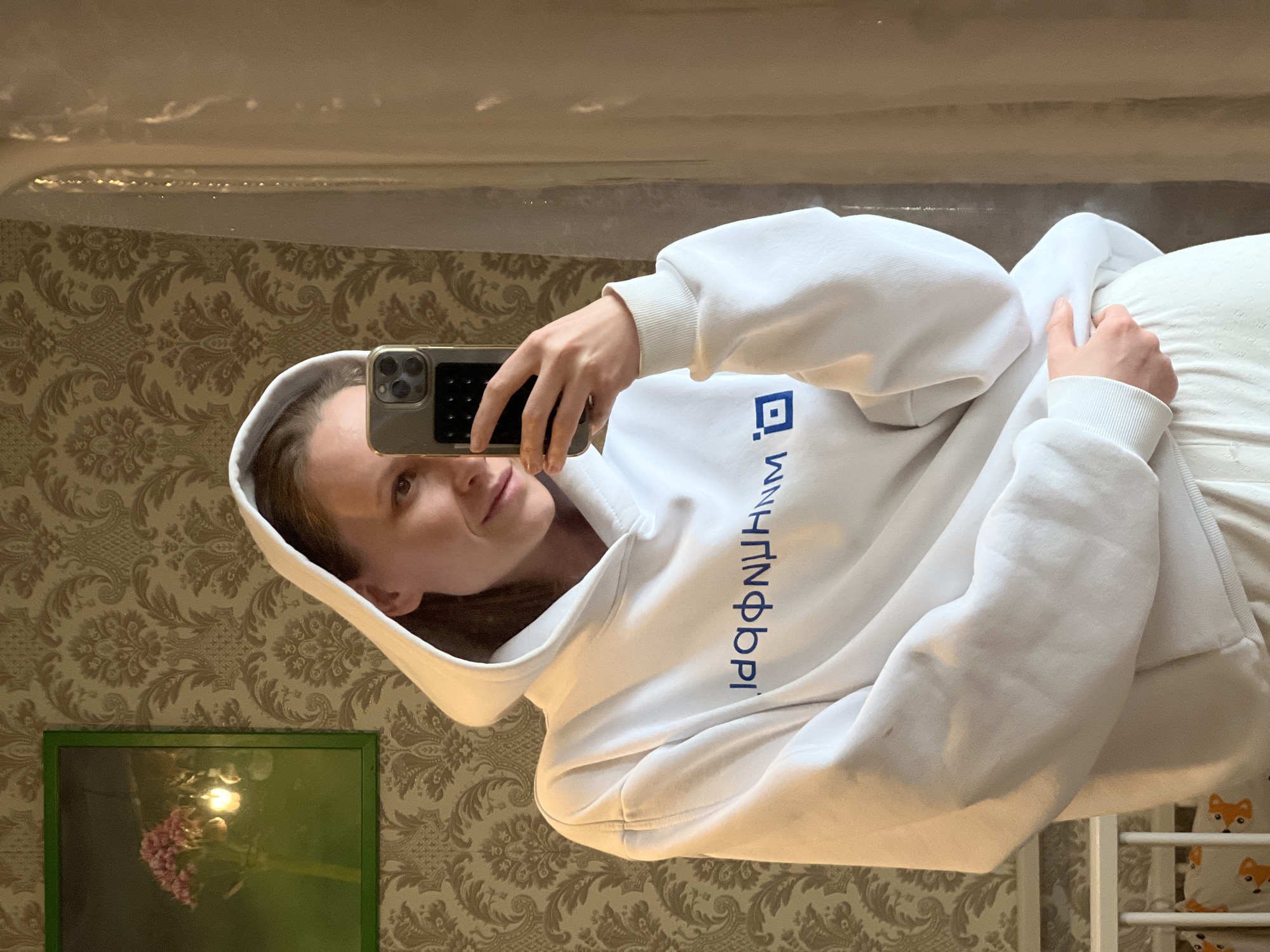 The image shows a young woman taking a mirror selfie indoors. She is positioned on the left side of the frame, holding a gold/khaki-colored iPhone with multiple cameras in her right hand to capture her reflection. She wears a white hoodie with a prominent blue square logo and blue Cyrillic text visible on the chest. The text appears to be "ДПИНФОРМ" (DPINFORM). Her face, seen in the mirror, has a neutral to slightly pleasant expression as she looks towards the phone.
The background, also reflected in the mirror, depicts an indoor room. A patterned wallpaper with a brown and gold damask-like design covers the wall. Below the mirror's edge, a green-framed picture or mirror reflects a light source and pink flowers. On the right side of the frame, a white bed or crib is partially visible, featuring a white sheet or pillow adorned with a pattern of orange and white fox heads. The setting suggests a personal space, likely a bedroom, in Razdolye, Russia.