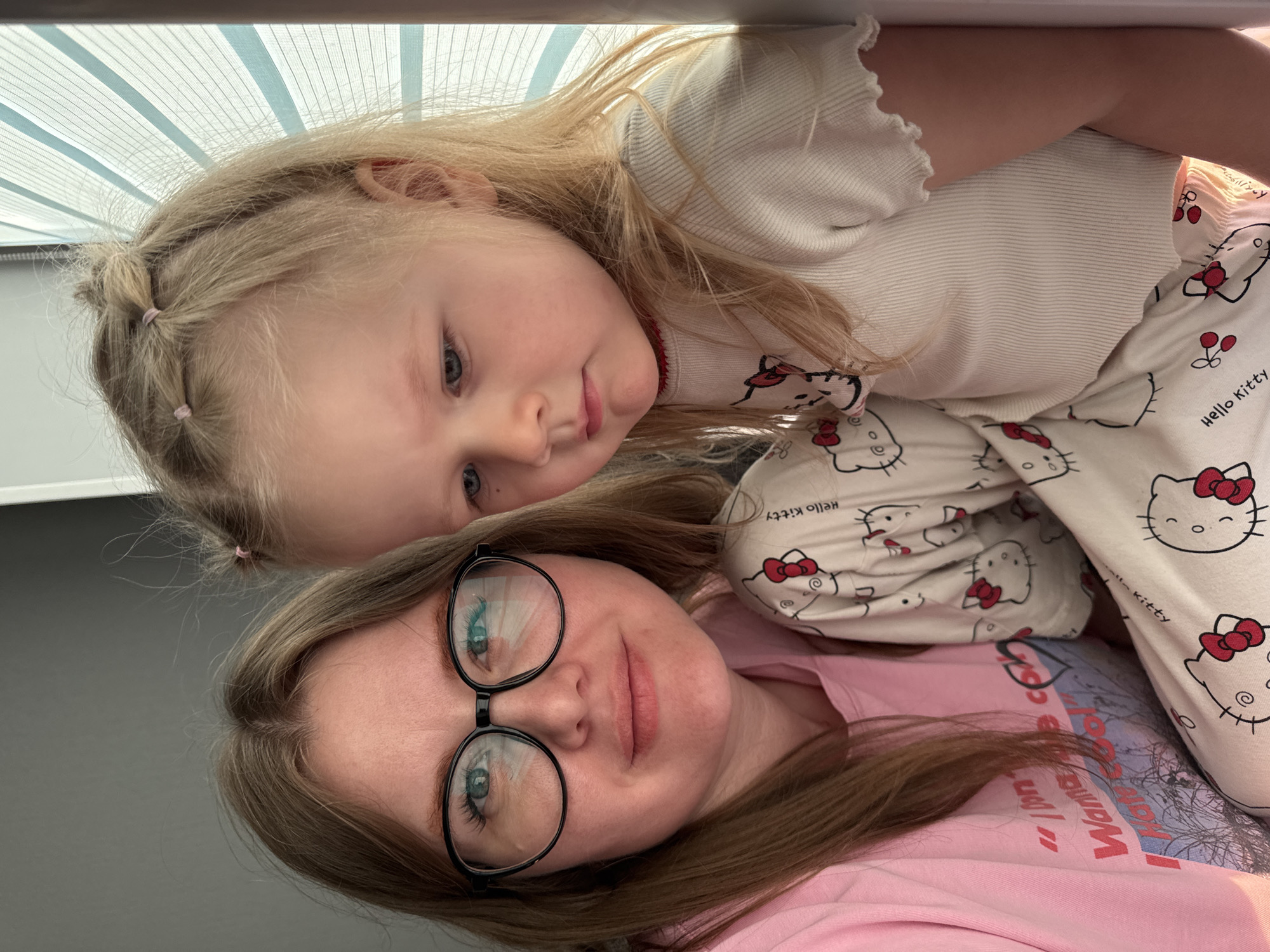 A woman and a young child are pictured indoors, possibly in a home. The woman is wearing glasses and a pink shirt, and the child is wearing pajamas with a "Hello Kitty" pattern. The child appears to be lying on or next to the woman. The background includes a window blind. The location is possibly in Gornyi (Dvurechenskoe S/P), Russia.