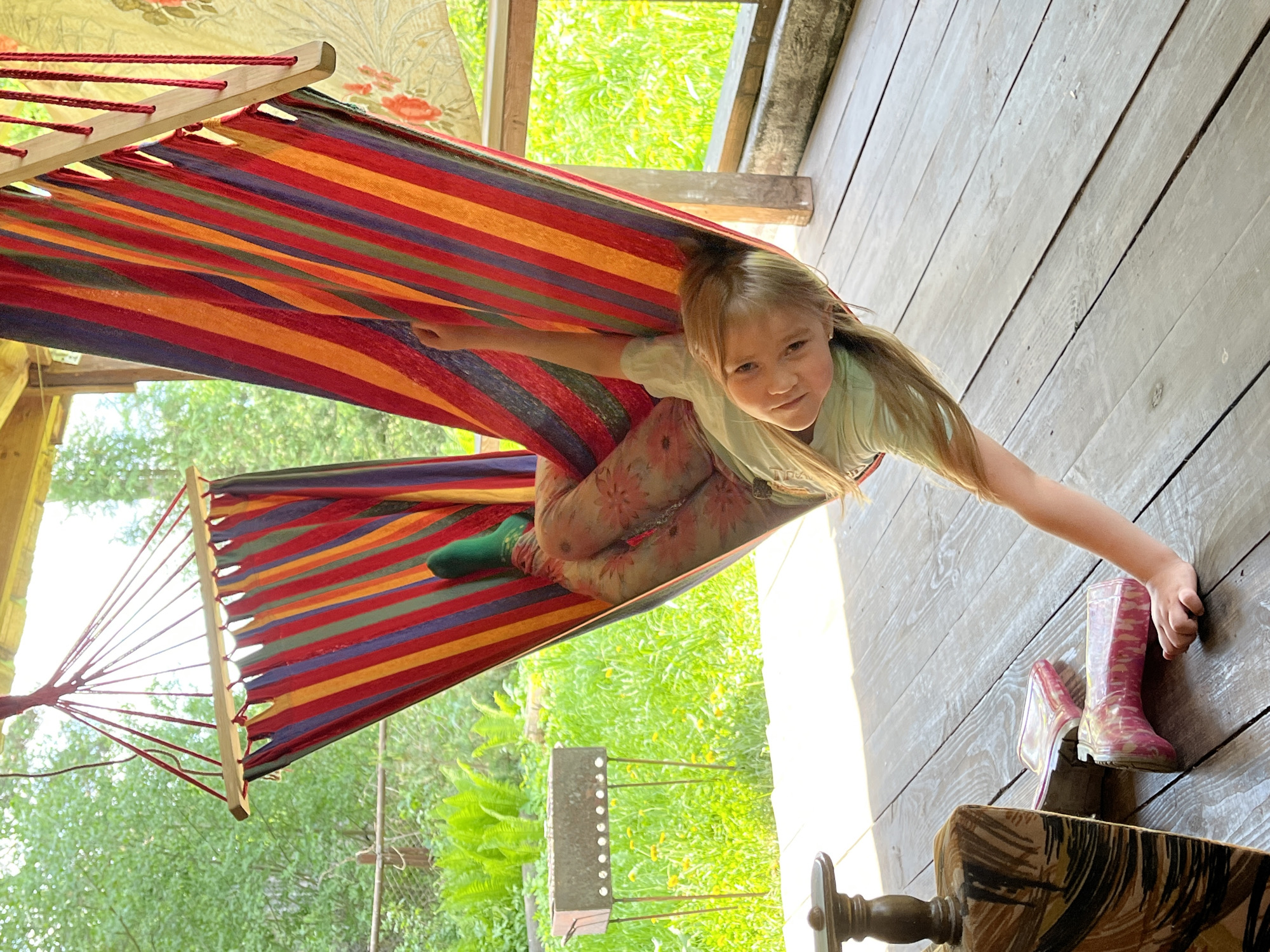 A young girl is lying in a multi-colored striped hammock. She wears patterned leggings and a light-colored shirt. One foot is covered in a green sock. One of her hands is reaching towards a pair of pink rain boots on a wooden deck. The hammock is set up outdoors, in what appears to be a garden or yard. The location is Chaikovsky, Russia.