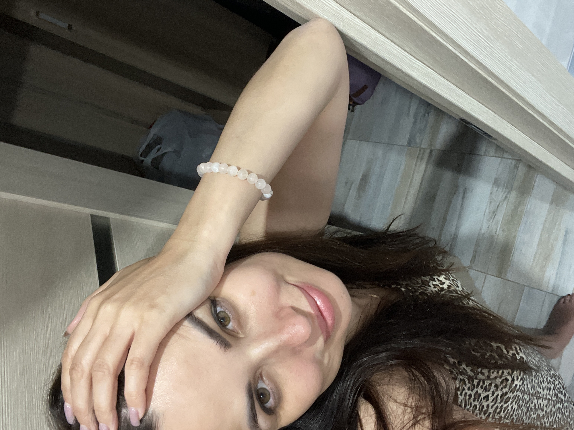 A woman with dark hair and brown eyes takes a selfie. She is wearing a leopard-print top and a pearl bracelet on her left wrist. Her left hand is resting on her forehead. The background appears to be a doorway with a closet visible behind her. The flooring is light-colored tile. The scene is indoors, in a domestic setting.