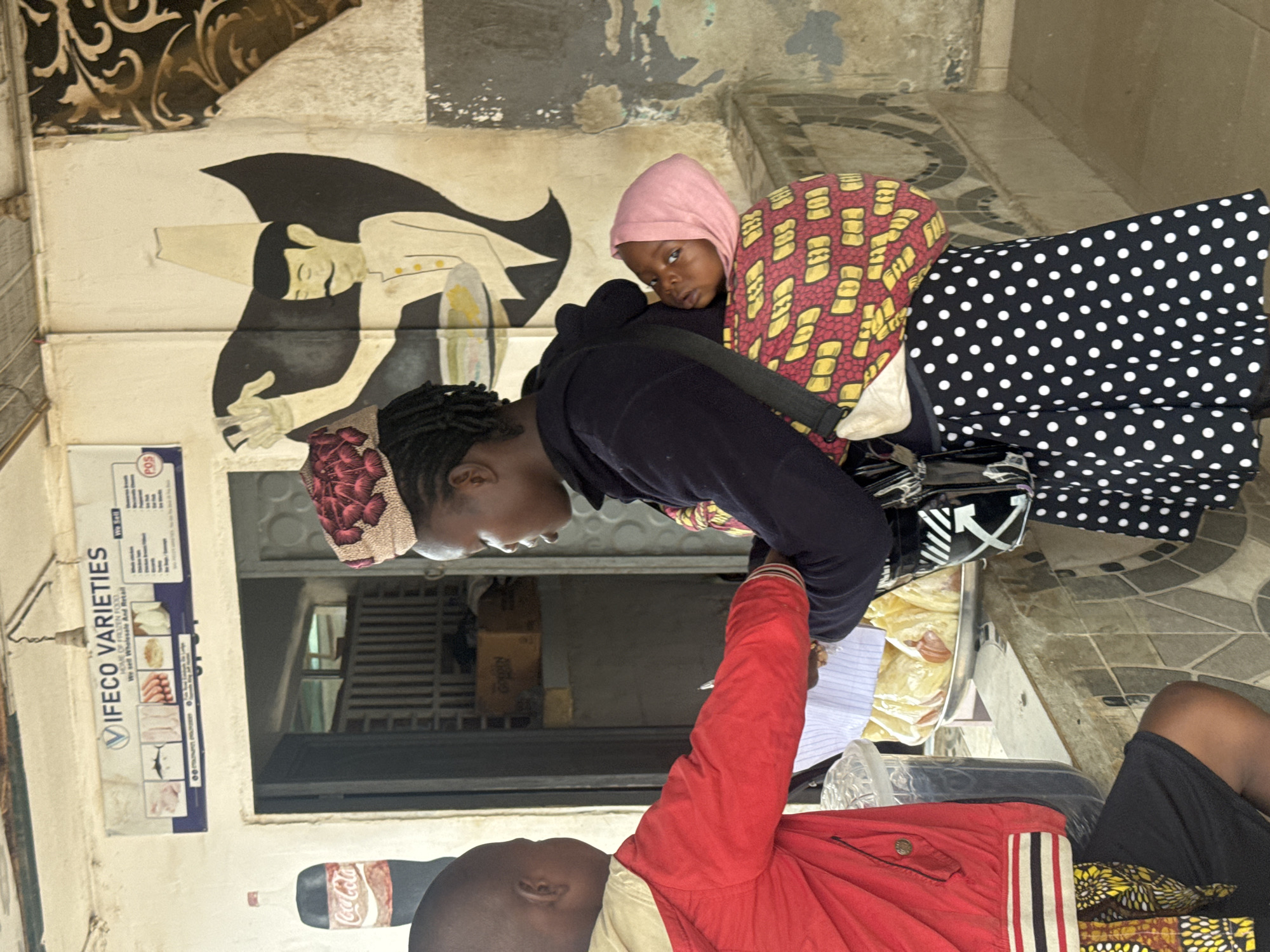 In Ihiagwa, Nigeria, a young boy in a red jacket stands facing a woman carrying a baby on her back. The boy is holding a piece of paper and appears to be engaged in a transaction with the woman. The woman is wearing a black hoodie and a patterned skirt, with a pink headscarf on the baby. Behind them, a store window is visible, with a sign that reads "IFECO VARIETIES. We sell Wholesale And Retail." A large painted mural of a man in a traditional hat is on the wall to the right. The scene appears to be an outdoor market or roadside shop.