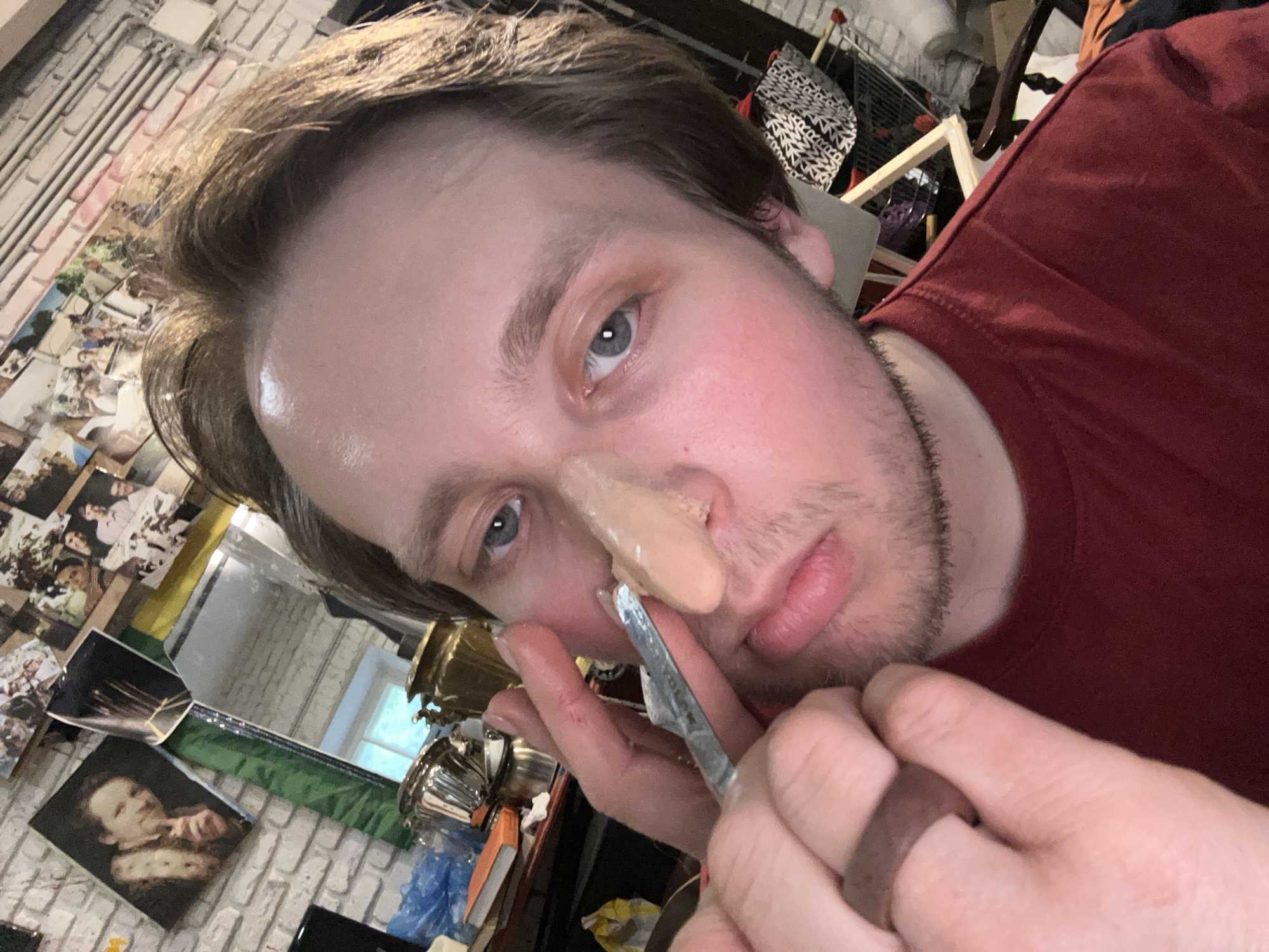 A person is applying a prosthetic nose. The person is fair-skinned with blue eyes, light brown hair, and some facial stubble. They are wearing a dark red t-shirt. The prosthetic nose is flesh-colored and appears to be in the process of being adhered to their nose with the aid of a metal spatula or tool held in their hand. The background shows a cluttered space with a wall covered in photos, assorted items on shelves, including metallic containers and a wooden frame. The location is indoors, and the city and area are unknown.