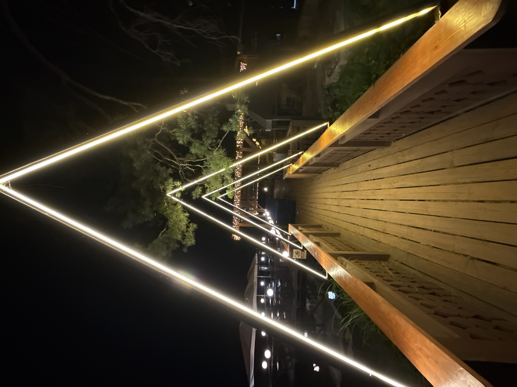 The image depicts a wooden boardwalk illuminated by triangular-shaped light installations. The boardwalk has wooden railings with decorative carvings. Beyond the boardwalk, there is a body of water with some structures and lights visible, suggesting a waterfront area or a resort. Trees with foliage are also present, framing the scene. The lighting suggests it is nighttime. The location is Zolotoi Plyazh, Russia.