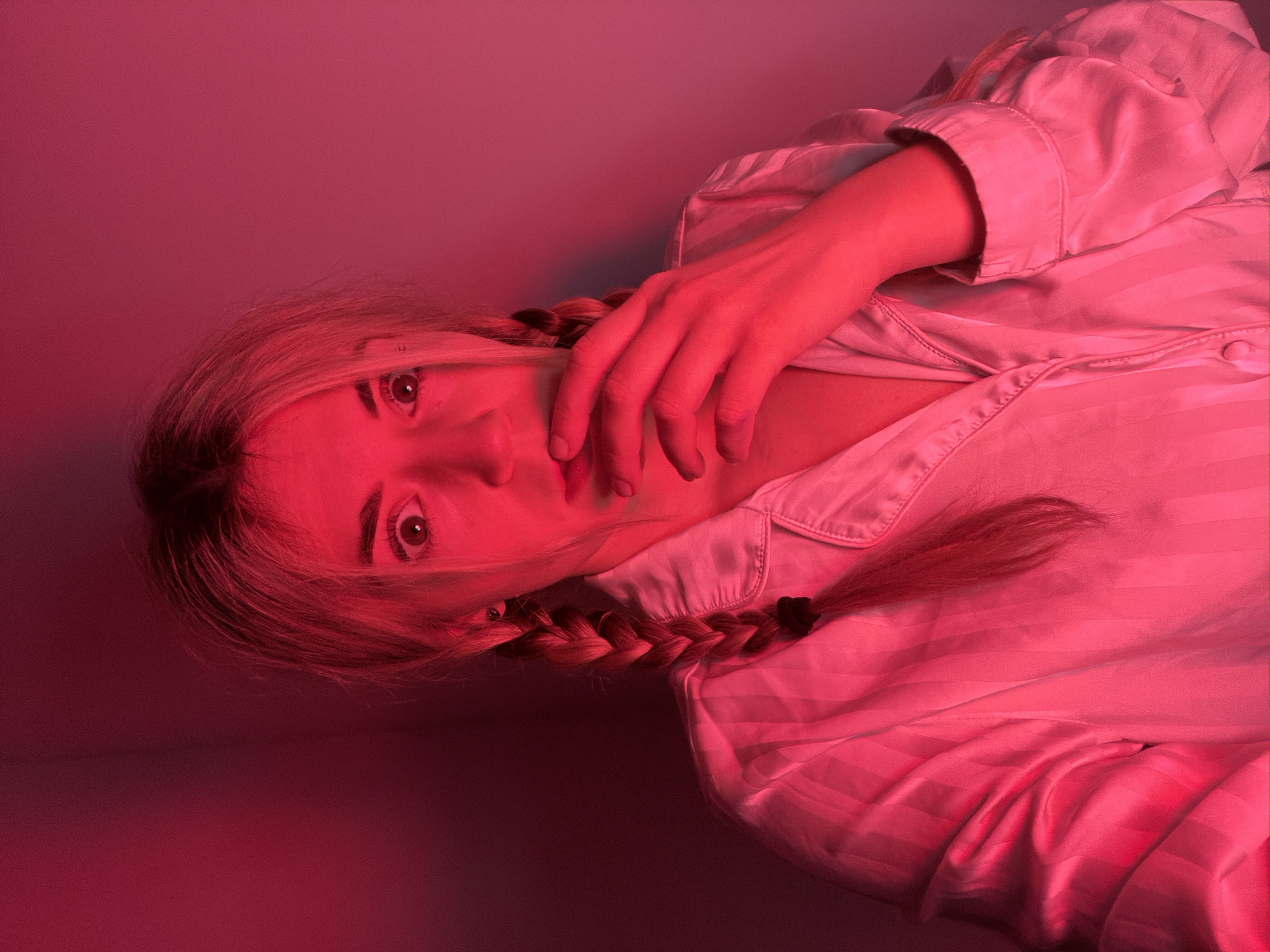 The image depicts a young adult female, illuminated by strong red light, positioned in an indoor setting. She has fair skin, wide-set eyes, and an open-mouthed expression of apparent surprise or shock, with her right hand partially covering her mouth. Her hair, which appears light-colored under the red illumination, is styled in two visible braids, one resting over her left shoulder. She is wearing a light-colored, possibly white or cream, collared top with vertical stripes, consistent with a pajama shirt. The background is a plain wall, uniformly saturated in red light. No other individuals or distinguishing objects are visible. The scene is located within an unspecified interior space in the city of Budva, Montenegro.