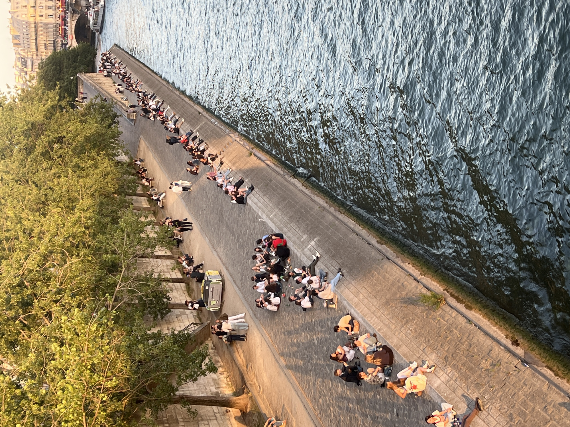 The image shows a long, stone embankment along the Seine River in Paris, France. Numerous people are seated and gathered on the embankment, enjoying the late afternoon sun. The trees lining the embankment are lush and green, casting shadows on the stone.
In the foreground, a group of people are sitting in a circle on the pavement. Some are holding bottles and appear to be socializing. Further up the embankment, more groups are dispersed, some sitting in rows along the edge of the river, others in smaller clusters. A few individuals are walking along the walkway.
The river itself is visible to the right, its surface rippled by a gentle current. In the background, a bridge spans the river, and buildings with characteristic Parisian architecture line the far bank. A boat is also visible on the river. The overall atmosphere appears relaxed and convivial, with people taking advantage of the pleasant weather and scenic location.