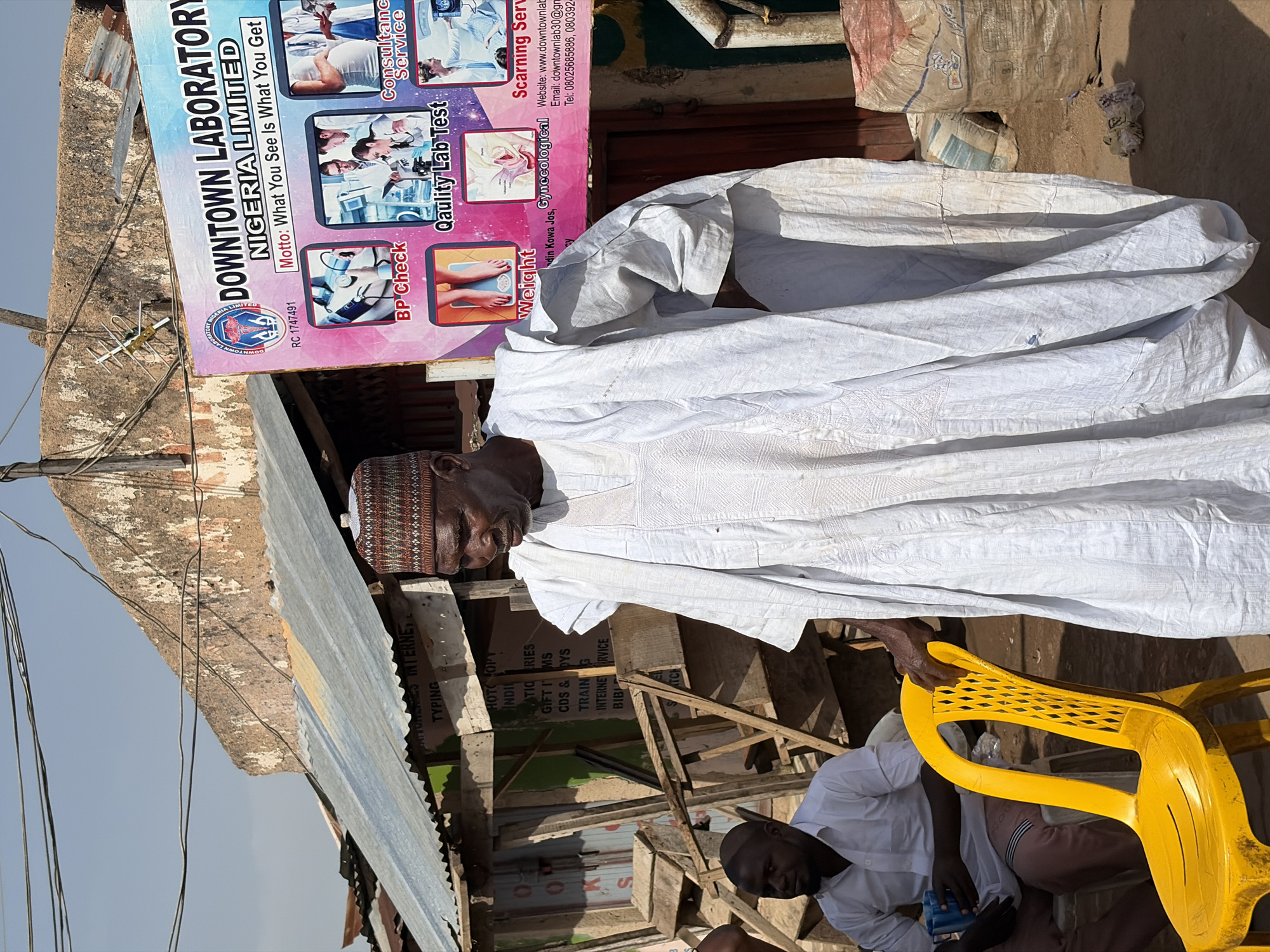 An outdoor scene features a man in traditional white clothing and a patterned kufi cap, standing beside a stack of large, draped white sacks. He holds a yellow plastic chair. Behind him is another person, also in a white shirt, sitting lower down. To the left is a weathered building, with a corrugated metal roof and wooden support beams. Signage visible on the building advertises "Downtown Laboratory Nigeria Limited," highlighting services such as BP checks, lab tests, weight management, consultation, and scanning. The location is indicated as being near Kowa Jos. The sign includes a website, email address, and telephone numbers. Additional hand-painted signs on the building advertise typing, internet, and other services. The surrounding environment appears to be a street setting under a bright sky.