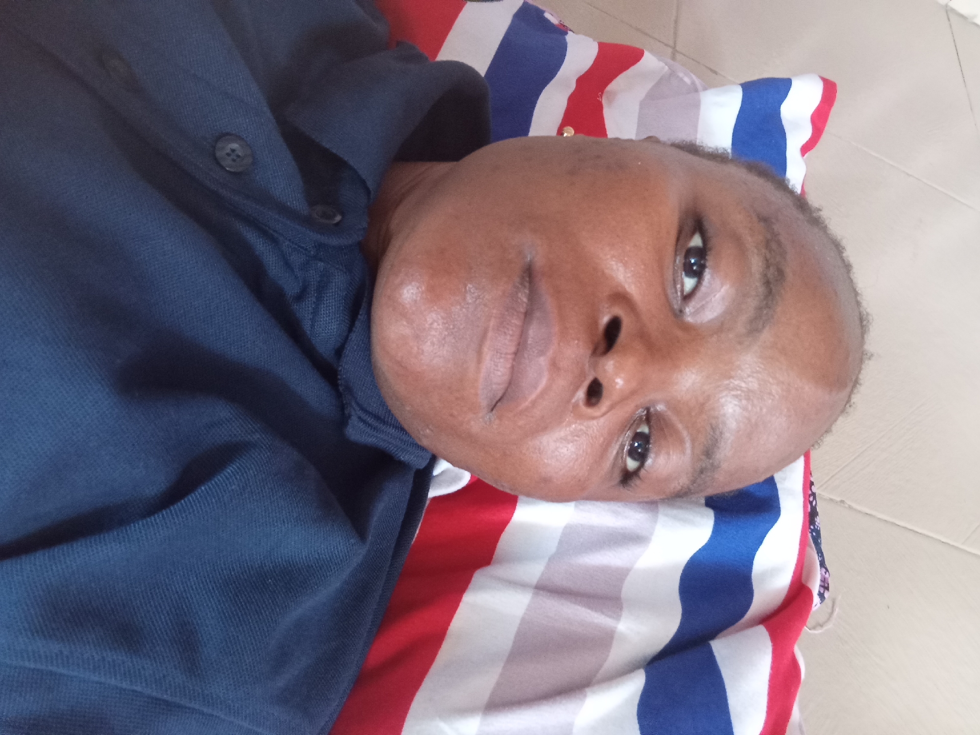 A close-up, overhead shot shows an adult with dark skin lying on a striped pillow in Kason, Nigeria. The individual is wearing a dark blue collared shirt. The pillow has bold red, white, and blue stripes. The person's face is turned slightly towards the camera, with their right eye visible and their left eye partially closed. Their skin appears smooth and slightly dewy. The background consists of a light-colored tiled floor.