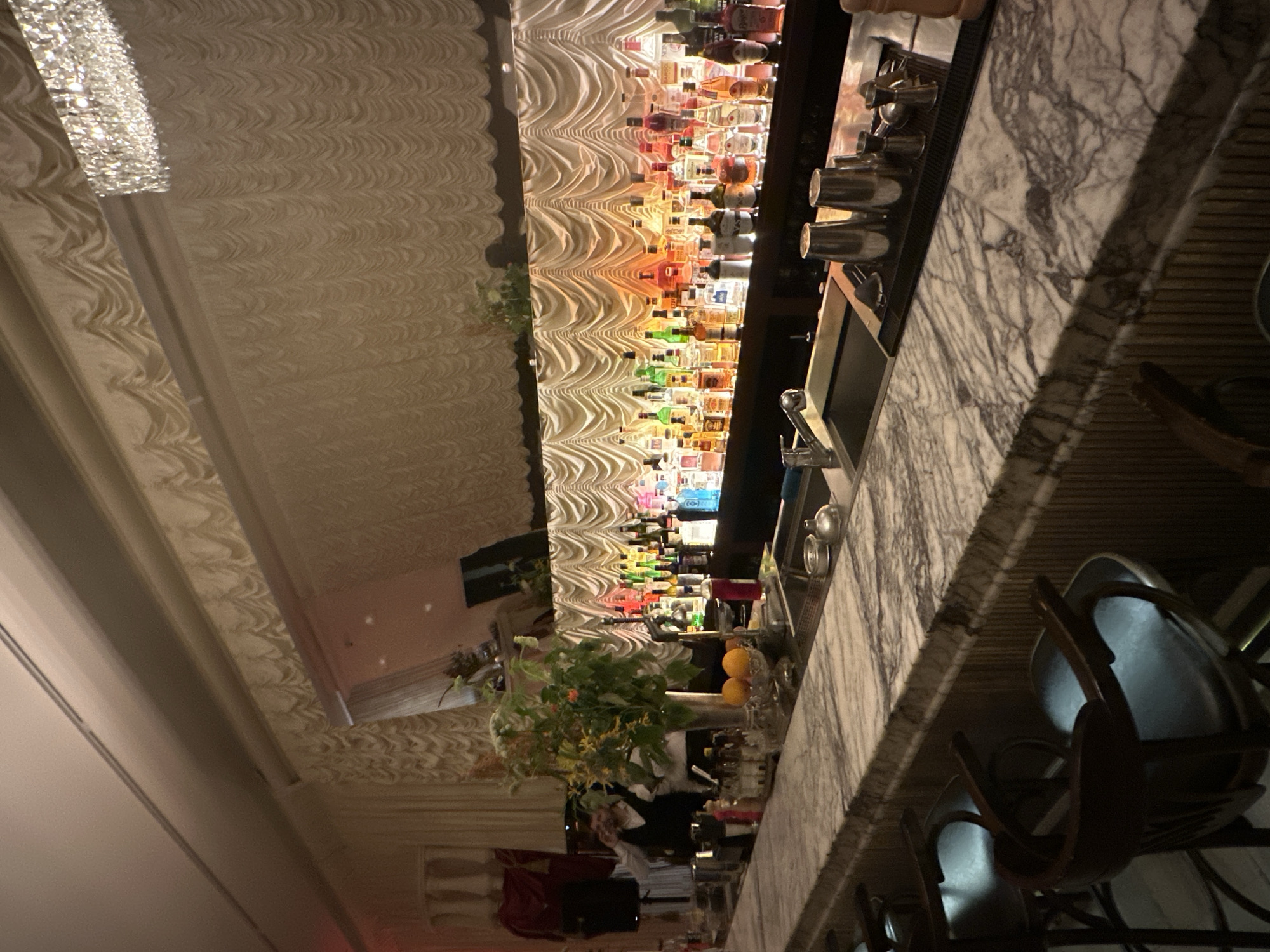 The image shows an indoor bar with a marble countertop and several bar stools. Behind the counter, a wall is lined with numerous bottles of alcohol, illuminated by colored lights. A chandelier hangs from the ceiling. A vase with flowers and two oranges are on the counter. A person is visible behind the bar. The scene does not provide any information about the city of Moscow, Russia.