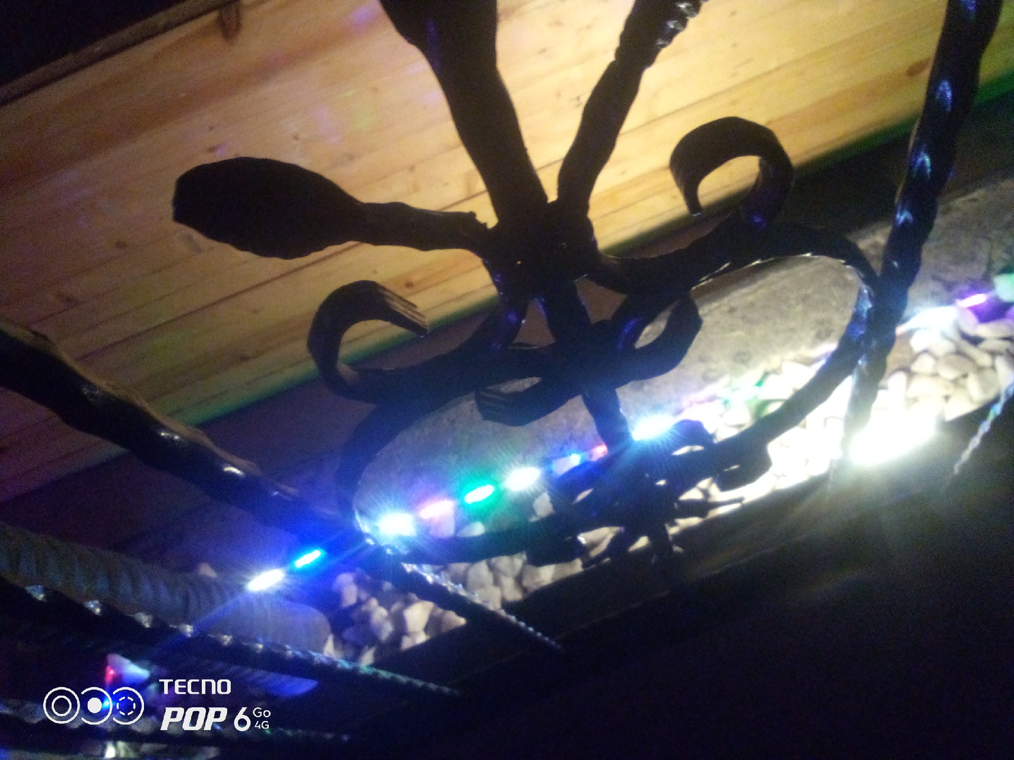 The image depicts a close-up, low-angle shot of a dark, intricate metal sculpture, possibly an artistic representation of an octopus or a similar multi-limbed creature. The sculpture is set against a backdrop of weathered wooden planks, illuminated by a string of small, colorful LED lights (blue, green, and white) that are wrapped around some of the sculpture's elements and extend downwards. The background also includes what appears to be gravel or small stones. The lighting is dim, with the LED lights providing the primary illumination, creating reflections and highlights on the metal and the surrounding surfaces. In the bottom left corner, a watermark indicates "TECNO POP 6 4G". The scene suggests an outdoor or semi-outdoor decorative setting, potentially in a garden or patio area. The specific location in Howayi, Nigeria, cannot be determined from the visual information provided.