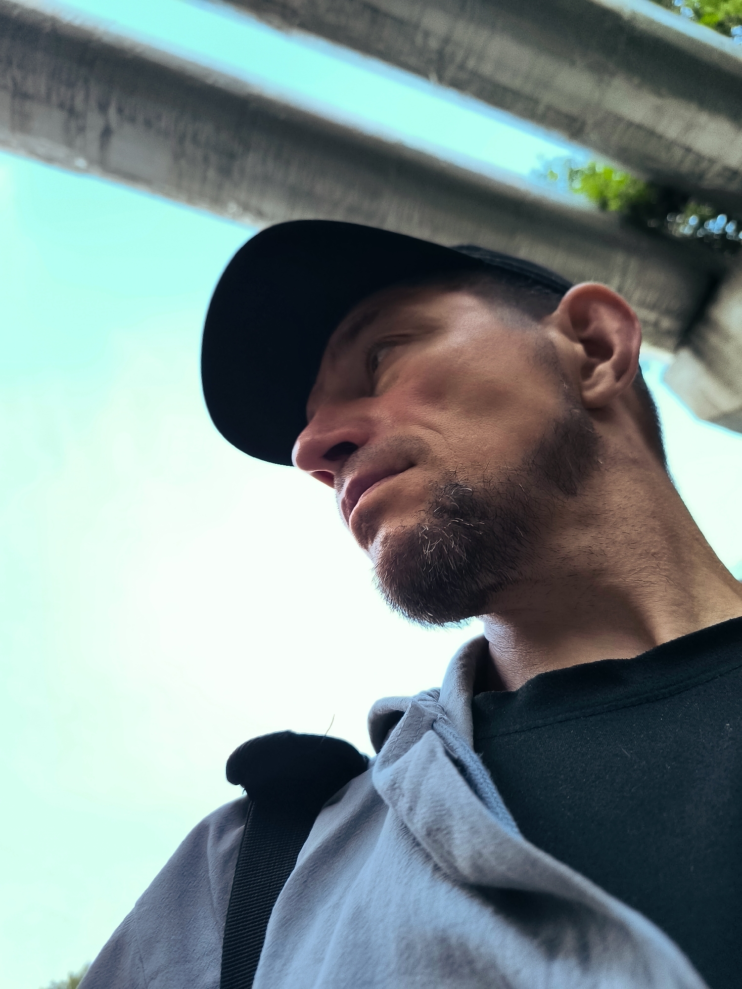 An extreme low angle, medium shot captures a man wearing a black baseball cap and a light grey hoodie over a dark t-shirt, looking downwards and to his left. He has short-cropped hair, stubble on his chin and jawline, and a prominent nose and ear. The background is a bright, clear sky with the underside of an overpass or similar concrete structure visible above. Green foliage peeks out from behind the concrete structure on the right side. The image is sharp and well-lit.