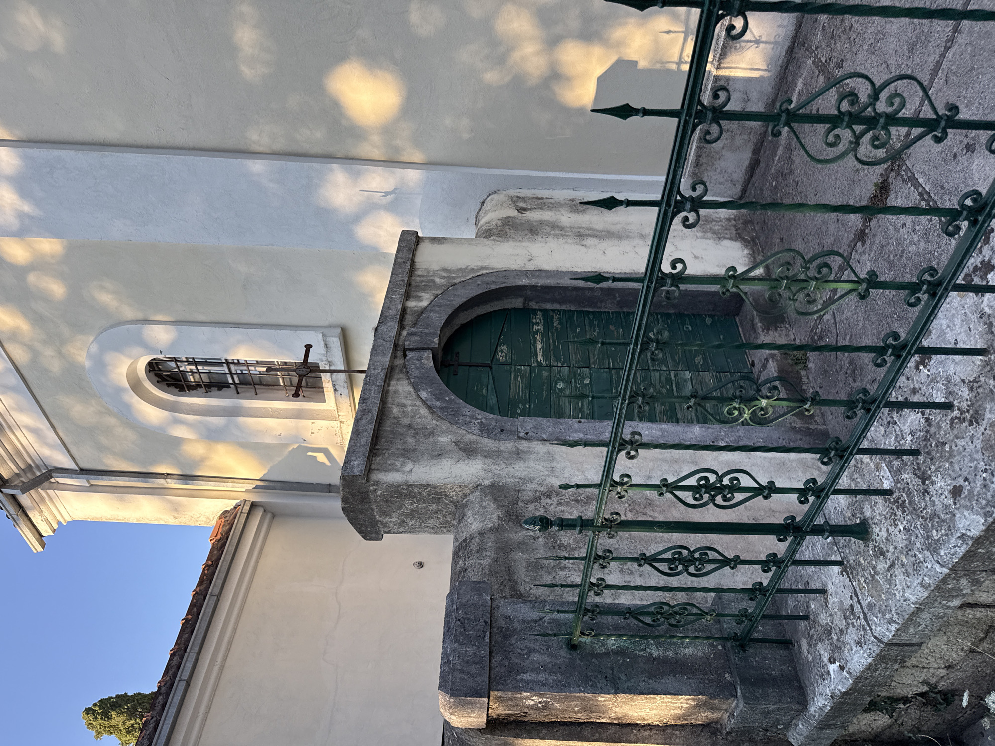 The image depicts the exterior of a building, possibly a church or a historical structure, in Đenovići, Montenegro. The building's facade is painted in a light cream color, with a prominent arched window featuring a metal grate. Sunlight casts dappled shadows across the wall, suggesting it is either morning or late afternoon. In front of the building, there is a stone wall with a green wrought-iron fence. Beyond the fence, a weathered green door is visible, set within a stone archway. The surrounding environment appears to be outdoors, with a clear blue sky visible at the top of the frame.