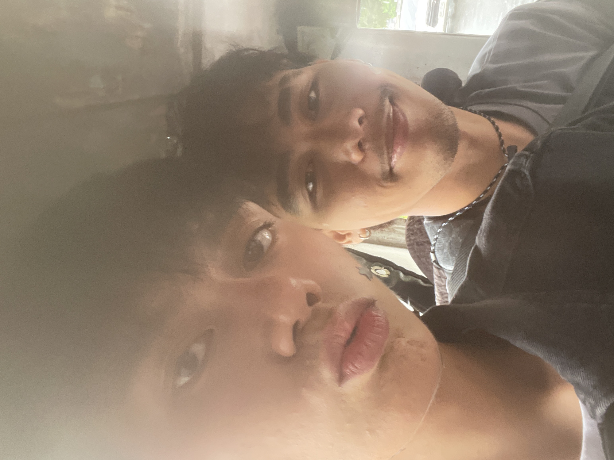 Two young men are captured in a close-up selfie, seemingly inside a vehicle. The man in the foreground, on the left, is leaning towards the camera, his face illuminated by soft light, revealing details of his lips and the left side of his face. He appears to be looking towards the camera. The second man, positioned to the right and slightly behind, is also looking towards the camera. He has a mustache and a subtle smile. Both men are wearing casual clothing. The background is blurred, suggesting the motion of the vehicle or a shallow depth of field, with hints of an urban environment, possibly Davao City, Philippines, due to the presence of a vehicle that resembles a tricycle, a common mode of transport in the region. The overall mood of the image is casual and personal.
