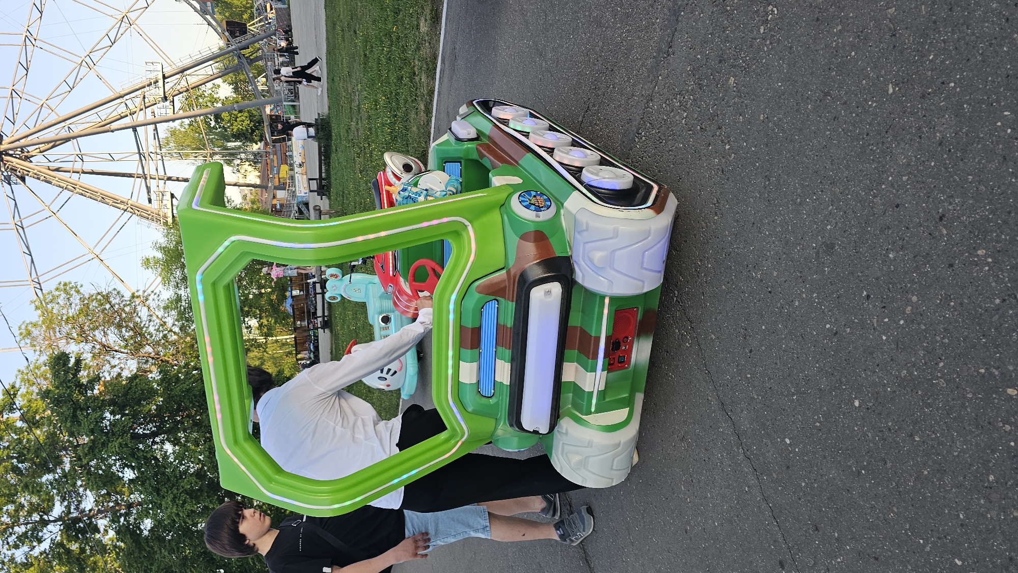 The image captures a scene at an amusement park. A green and camouflage-patterned ride, possibly designed to resemble a vehicle, stands on an asphalt surface with a crack running across it. Two individuals are positioned near the ride. One, wearing a white long-sleeved shirt and black pants, appears to be interacting with the ride. Next to them is another person in a black t-shirt, shorts, and sneakers. In the background, a large Ferris wheel is visible against a blue sky, indicating an outdoor setting. Green grass and trees are also present, typical of a park or fairground environment.