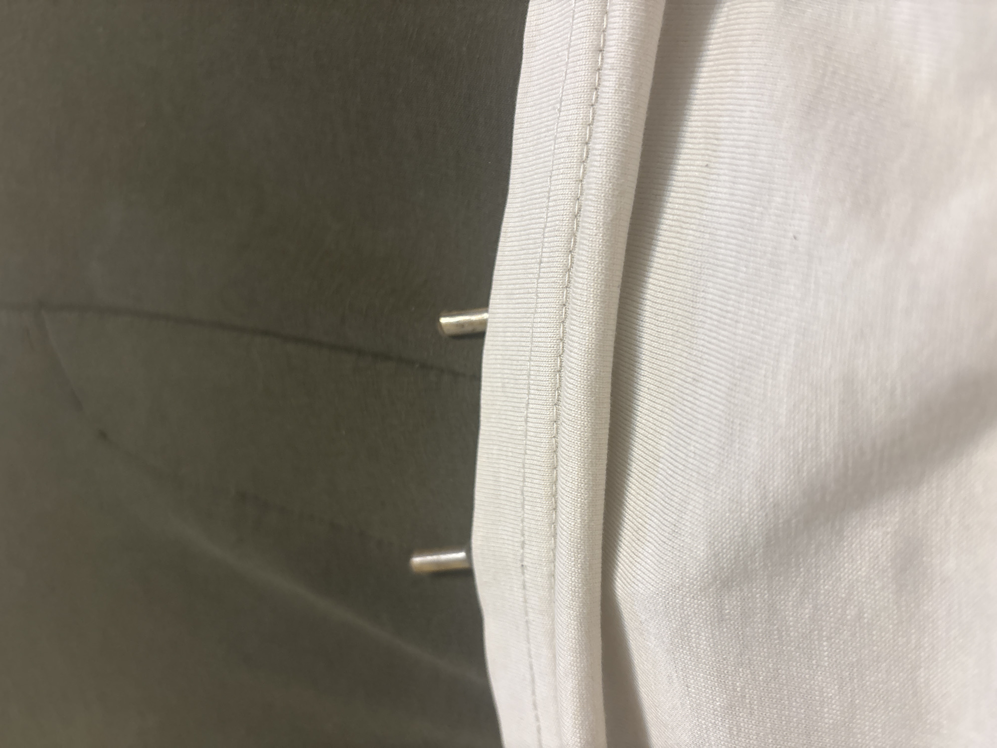 The image shows a close-up view of two metal aglets, or the tips of a drawstring, protruding from a white fabric. The aglets are cylindrical and silver in color. They are positioned on the edge of a garment, likely a hoodie or sweatpants, where a drawstring would typically be threaded through. The white fabric appears to be ribbed or textured. In the background, a darker, olive-green fabric is visible, possibly part of the same garment or another item of clothing. The lighting suggests an indoor setting, with no discernible city or location details present.