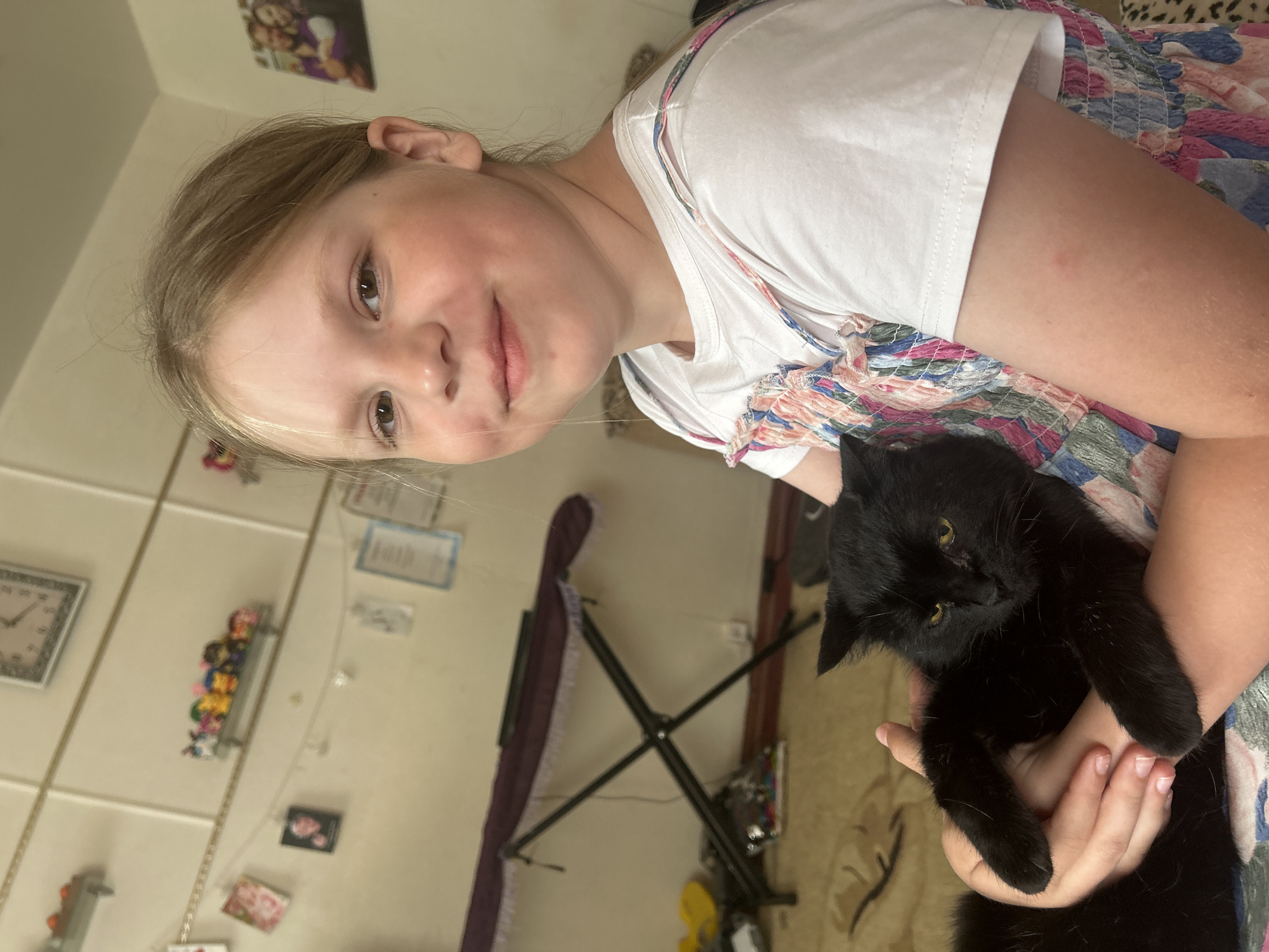 A young girl holds a black cat. The girl has blonde hair and is wearing a white t-shirt and a floral dress. The black cat is cradled in her arms. The background is a room with a clock, shelves, and a music stand. It is not possible to ascertain the location within Фоки, Russia.