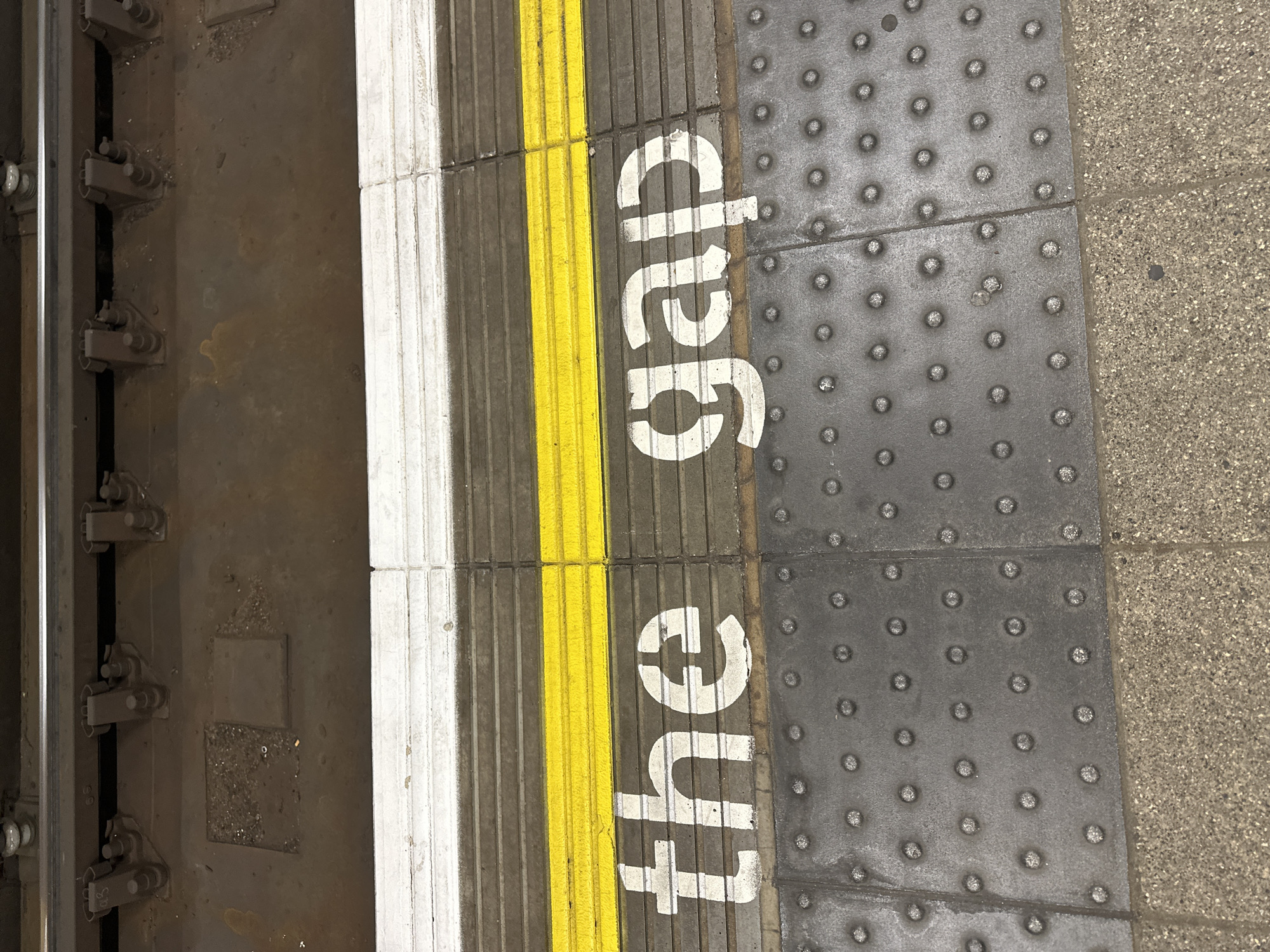 The image captures a close-up, downward-angled view of a train station platform edge, strongly suggesting a London Underground or National Rail station. On the far left, a section of a dark metal railway track is visible, with connecting components and fasteners.
Adjacent to the track is the platform edge, featuring distinct safety markings and tactile paving. A narrow, off-white stripe runs parallel to the track, followed by a prominent yellow corrugated strip designed for visual and tactile warnings. Immediately to the right of the yellow strip, on a darker grey, ribbed section of the platform, the white stenciled words "the gap" are clearly visible, oriented vertically. This phrase is a well-known warning to passengers about the space between the train and the platform.
Further to the right, a section of dark grey tactile paving with raised circular studs is present, intended to assist visually impaired passengers. This studded paving abuts a lighter, rough-textured concrete or stone surface that makes up the main platform area. The surfaces show signs of wear and some grime, consistent with a heavily used public transport environment. The lighting is even, suggesting an overhead light source or diffused daylight. No people or active events are captured in the frame, focusing entirely on the platform infrastructure.