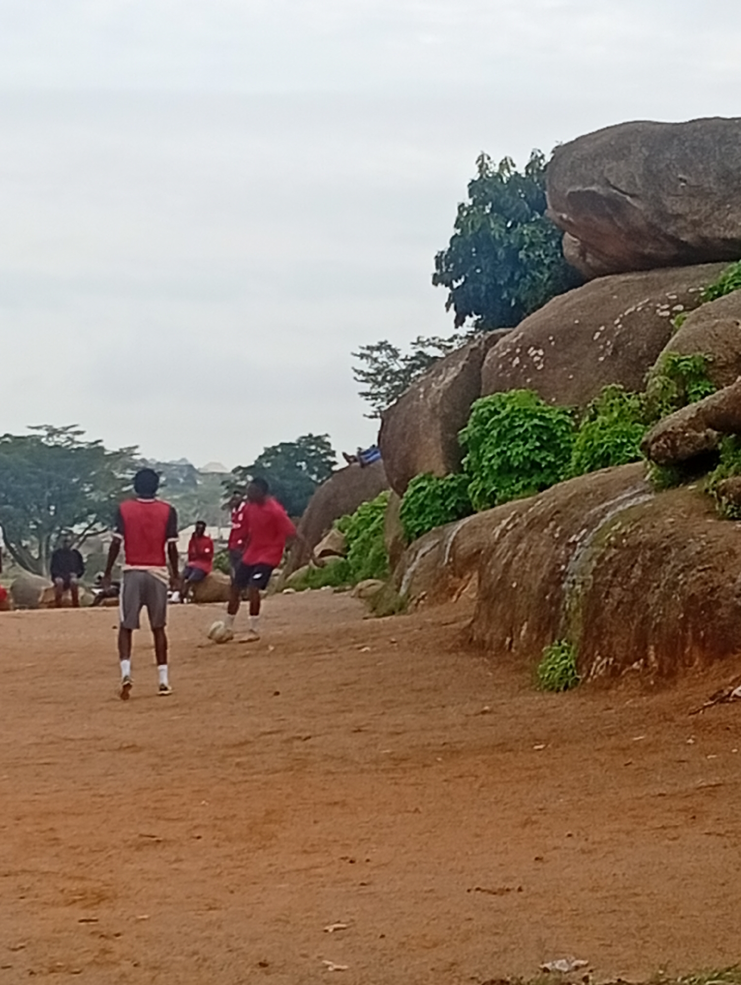 The image depicts an outdoor scene in Jos, Nigeria, featuring a group of young men playing football on a dirt field. The foreground is dominated by the sandy terrain, with scattered small rocks and debris. Two individuals in red shirts and grey shorts are actively engaged in the game, with one in the process of kicking a white football. In the background, several other people are visible, some seated and others standing, observing the match. The environment is characterized by large rock formations and sparse vegetation, typical of the rocky landscape found in Jos. The sky is overcast, suggesting a cloudy day. The overall atmosphere appears casual and recreational.