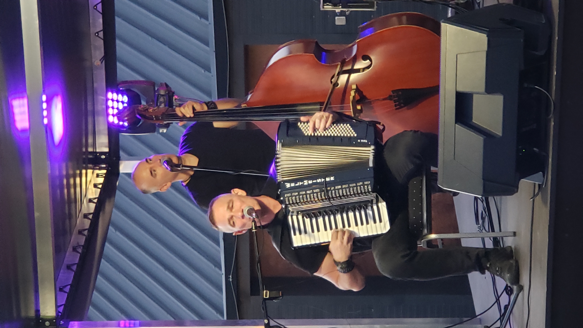 Two men are performing music on a stage at night in Prešov, Slovakia. The man in the foreground, seated on a metal chair, plays an accordion and sings into a microphone. He wears a black short-sleeved shirt and black pants. Behind him, to the right, a second man stands and plays a double bass. He is wearing a black t-shirt. The stage is illuminated by purple stage lights overhead, and there are speakers in the foreground. The background consists of a slatted wall.