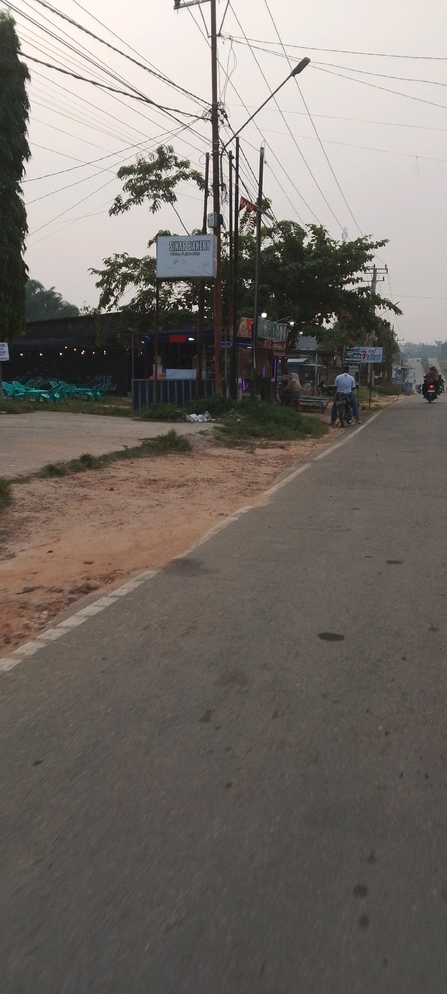 The image depicts a street scene in Lenggadai Hulu, Indonesia, during what appears to be late afternoon or early evening. The perspective is from a moving vehicle, looking towards a roadside area with shops and parked motorcycles. A prominent sign for "SINAR BAKERY" is visible, indicating a local business. Several individuals are present, some sitting outside a shop and one man on a motorcycle, possibly waiting. Green plastic chairs and tables are set up outside a dark-colored establishment, suggesting an outdoor seating area. A dirt path runs alongside the road, and overhead power lines are abundant, a common feature in many Indonesian towns. In the distance, the road continues, with a few more people on motorcycles visible further down. The overall impression is of a quiet, everyday scene in a rural or semi-urban Indonesian setting.