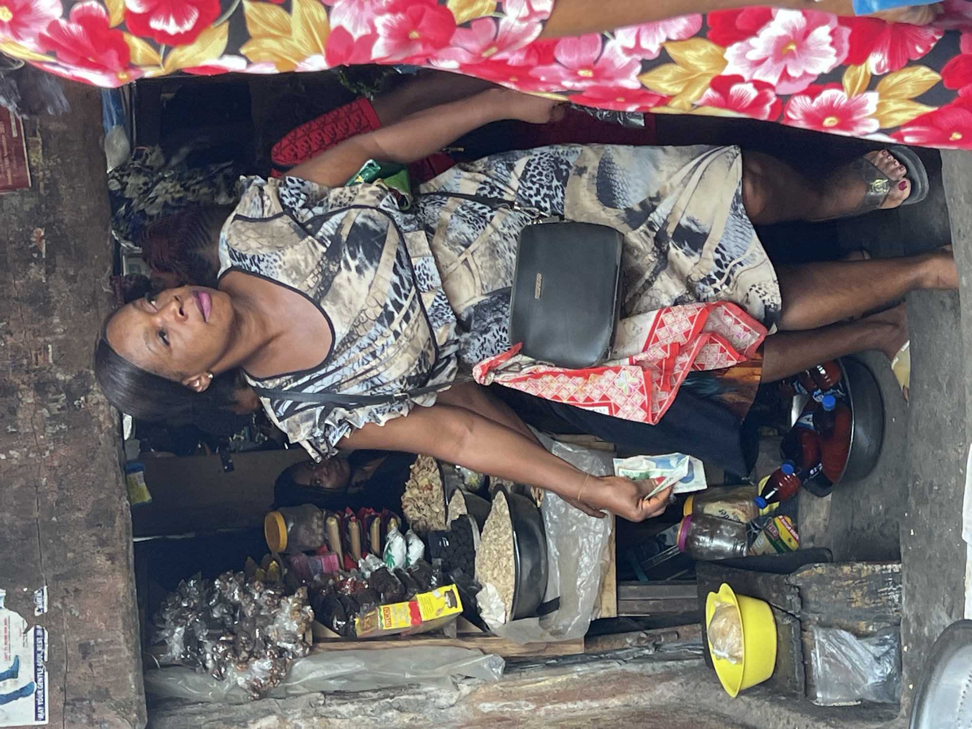 The image, taken in Nkpor, Nigeria, depicts a woman attending a small market stall or shop, likely during the daytime. The perspective is from slightly above and angled.
The primary subject is a woman with dark hair, wearing a patterned dress featuring tan, black, and white animal print or abstract designs. She has light pink lipstick and a neutral expression, looking slightly upwards and to her left. A black handbag or wallet rests on her lap. In her left hand, she holds several banknotes, suggesting a transaction. Her right leg and foot, wearing a clear-strapped sandal, are visible. Above her, another person's arm and leg are partially visible, clad in a vibrant floral fabric with red, yellow, and white flowers on a dark background. Another individual's head and shoulders are dimly visible in the background, behind the main woman.
The stall itself is rustic, with rough, possibly concrete or mud-brick walls. Merchandise is displayed on makeshift wooden shelves and directly on the ground. Visible items for sale include large metal bowls filled with what appear to be dried beans, nuts, or seeds. Another bowl contains multiple bottles with reddish liquid, possibly palm oil or drinks. Various packaged goods are scattered, some in clear plastic bags, and a yellow packet labeled "MAGGI" is clearly visible. A yellow plastic bowl holds a wrapped item. The overall environment suggests an informal, bustling market setting. On the far left, a poster is partially visible on the wall, with faint text including "MAY YOUR GENTLE SOUL REST IN."