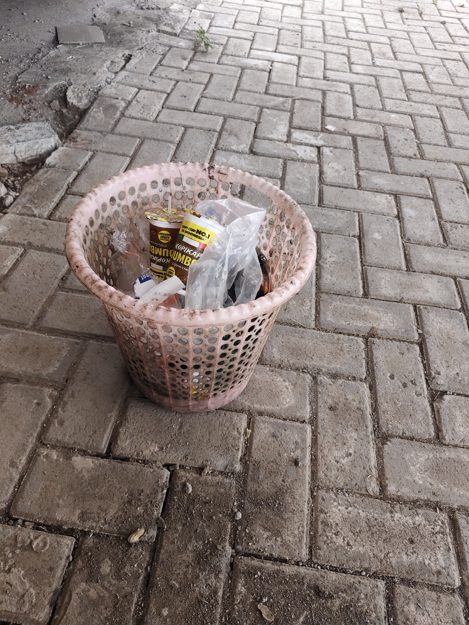 The image shows a pink plastic trash can filled with refuse. The trash can is situated on a paved area made of interlocking brick pavers. The pavers are weathered and show signs of dirt and wear. In the background, to the left, there is a patch of rough, unpaved ground with some debris. To the right of the trash can, the pavers continue, with a small patch of green vegetation growing between some of the bricks. The contents of the trash can include plastic bags, paper cups, and other assorted waste. One of the paper cups visible is labeled "KOPI CUP NO.1 DI INDONESIA". The lighting suggests it is daytime, with natural light illuminating the scene. The overall environment appears to be an outdoor urban or semi-urban setting in Bangsalsari, Indonesia. There are no people visible in the image.