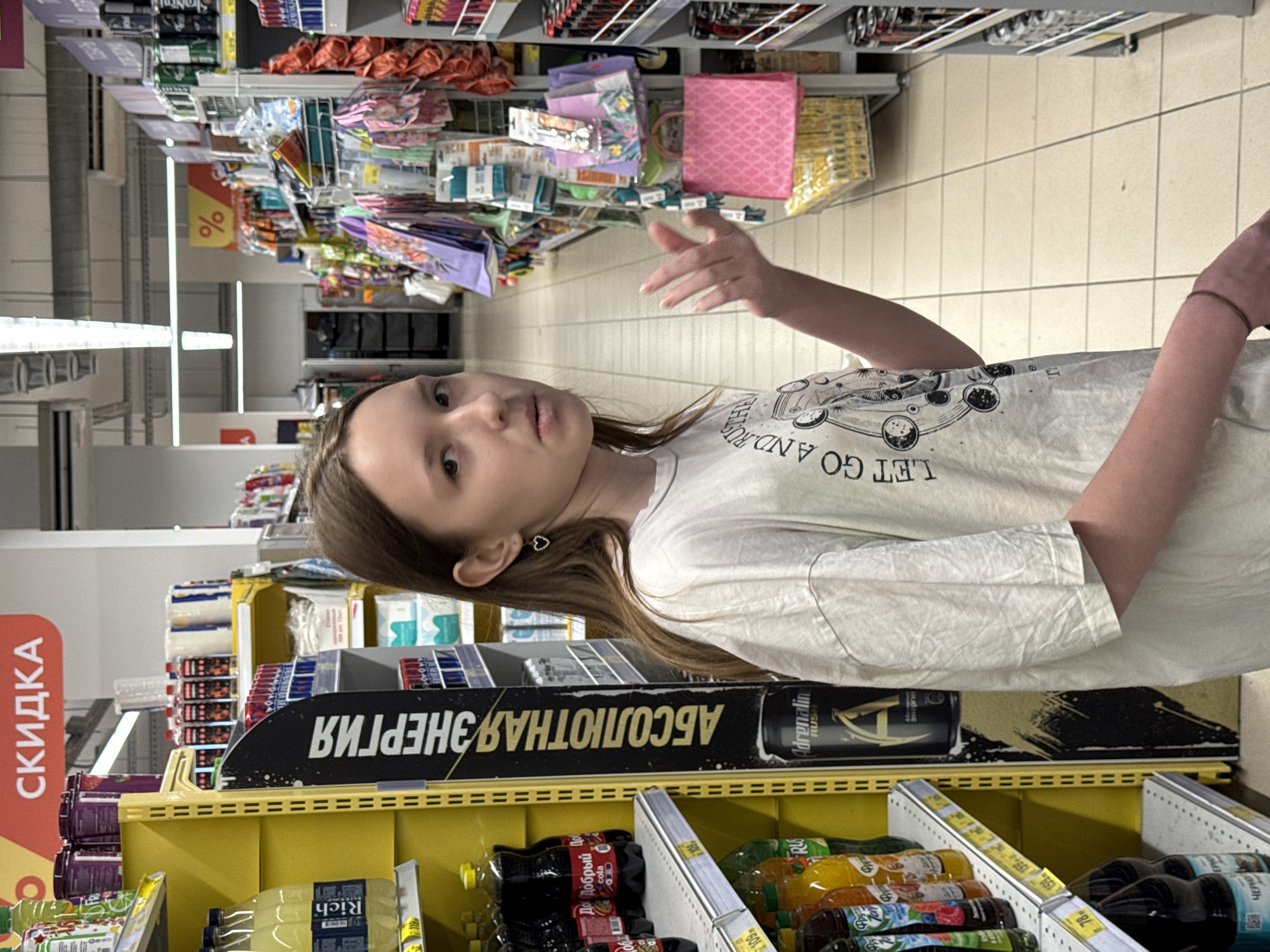 A girl stands in the aisle of a supermarket, likely in Rostov-on-Don, Russia, based on the Cyrillic script on the product labels and the "Adrenalin Rush" energy drink brand. She is wearing a white t-shirt with the text "LET GO AND RUS" and gestures with her right hand. Behind her are shelves stocked with various items, including energy drinks, juices, and other products. A black display banner promoting "ABSOLUTE ENERGY" is visible in front of the shelves. Sale signs in Russian ("СКИДКА") are also present. The background shows more supermarket aisles and products.