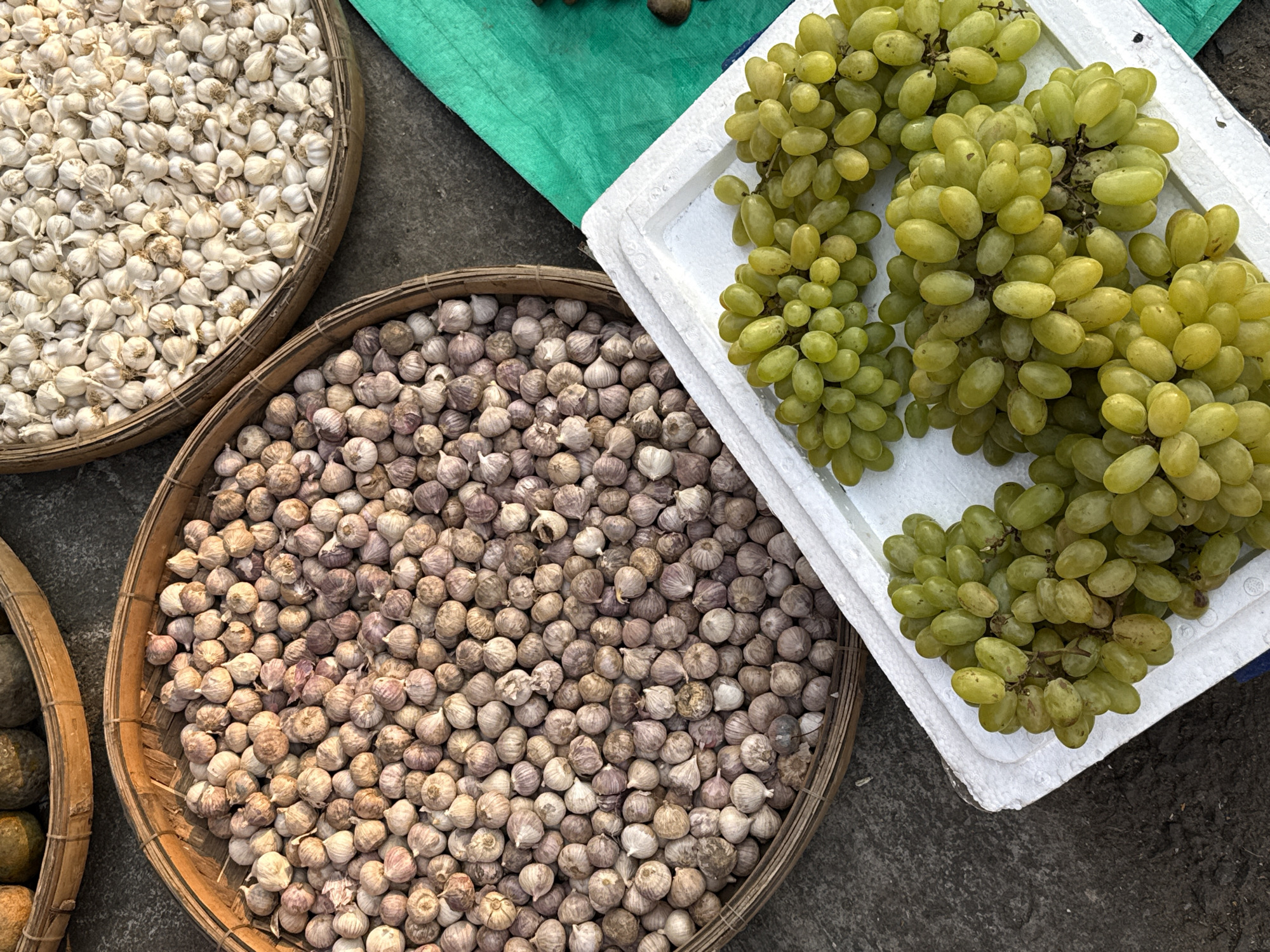 An outdoor market stall in Bắc Nha Trang, Vietnam, displays produce laid out for sale on a rough, grey surface. To the left are two circular, woven baskets. One holds small, whitish garlic bulbs, while the other contains slightly larger, purple-tinged garlic bulbs. To the right, green grapes are arranged on a white styrofoam tray. A small corner of green cloth is visible at the top.