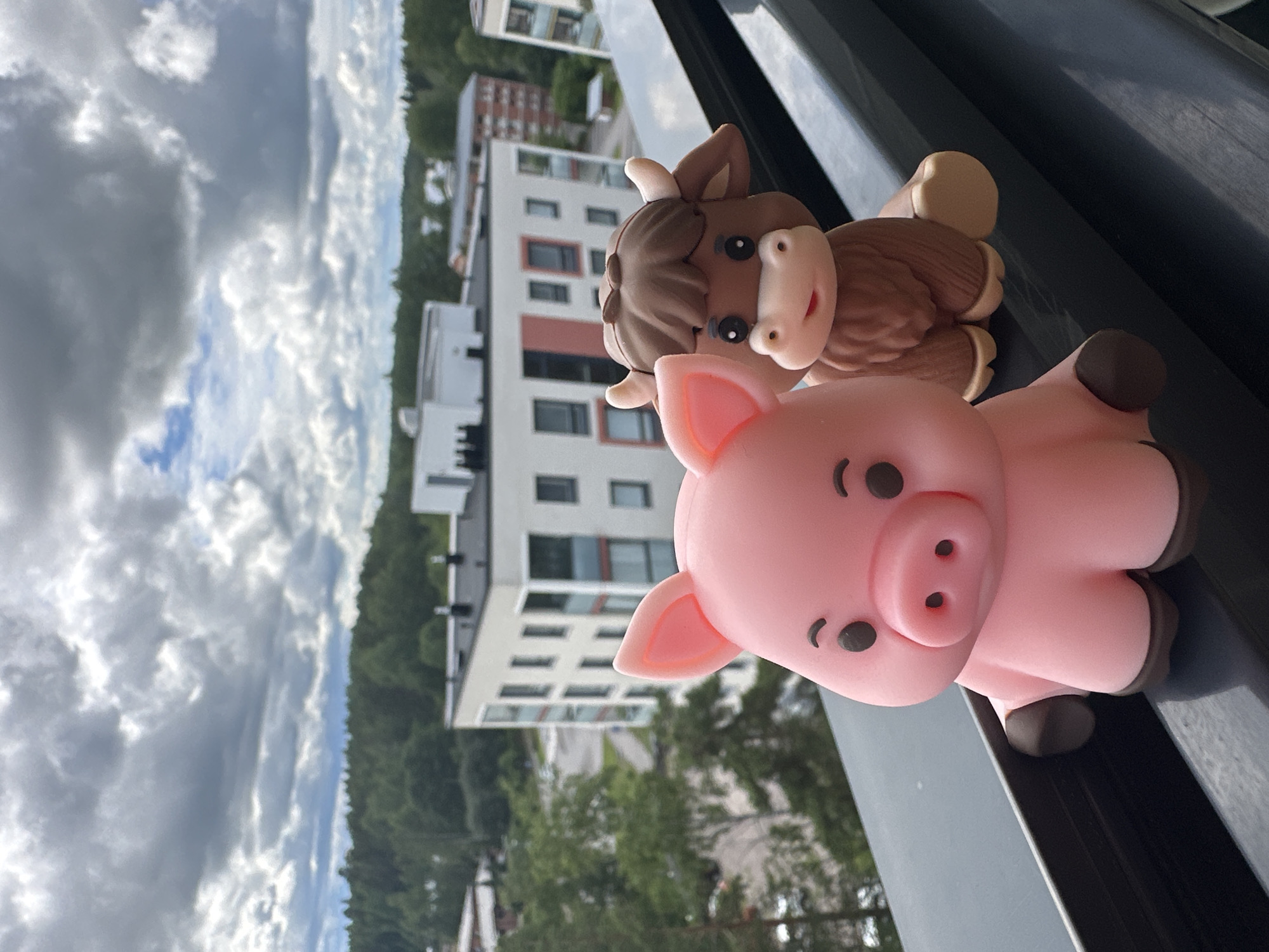 Two small, stylized figurines, a pink pig and a brown Highland cow, are positioned on a dark ledge in the foreground. The pig is to the left, facing forward with its snout prominent. The cow is to the right, slightly behind the pig, also facing forward. In the background, a blurred urban landscape is visible, featuring modern apartment buildings surrounded by green trees. The sky above is overcast with dramatic, dark clouds.