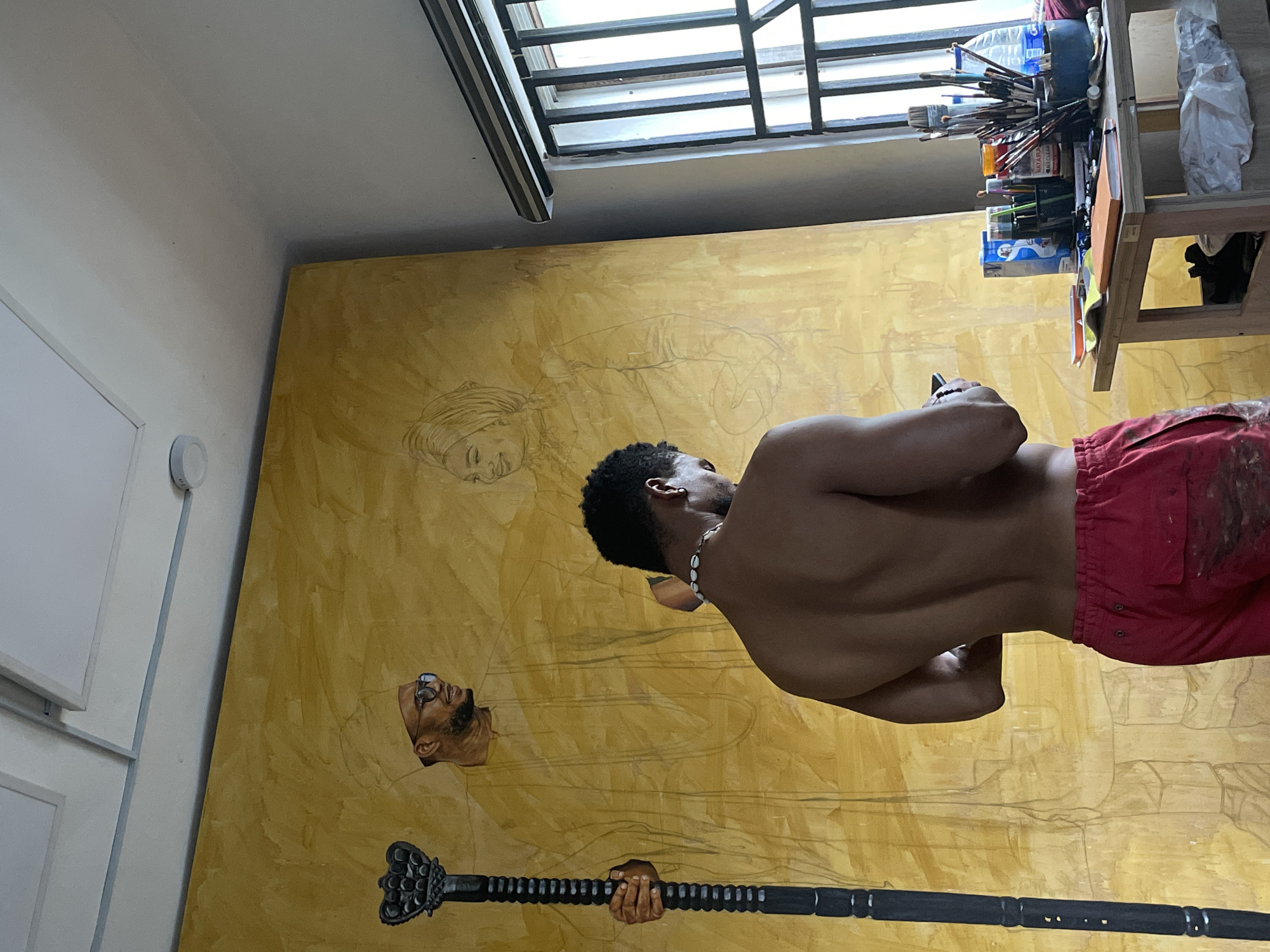 The image captures an artist's studio, likely in Ado, Nigeria, centered around a large, golden-toned canvas on a wall. A shirtless man, seen from behind, stands before the artwork, engrossed in a black smartphone he holds. He has short, dark, curly hair, wears a white shell necklace, and red shorts stained with paint.
The large canvas displays an intricate painting in progress. A prominent, realistically rendered head of a bearded man with glasses, looking upwards, is visible on the lower left. Below this, a painted hand firmly grips a dark, ornate staff or scepter. The rest of the golden background features faint pencil outlines of other figures, including a less defined face, possibly a woman's, situated above and to the left of the bearded man's head.
To the right, a wooden shelving unit is filled with various art supplies, such as numerous paintbrushes, tubes, and bottles. Text like "MATAR" and "CLEAN" can be partially discerned on some of the packaging. A large window with horizontal bars at the top of the frame suggests ample natural light. To the left, a white wall with a circular smoke detector completes the indoor studio setting. The artist appears to be taking a break from painting, checking his phone.