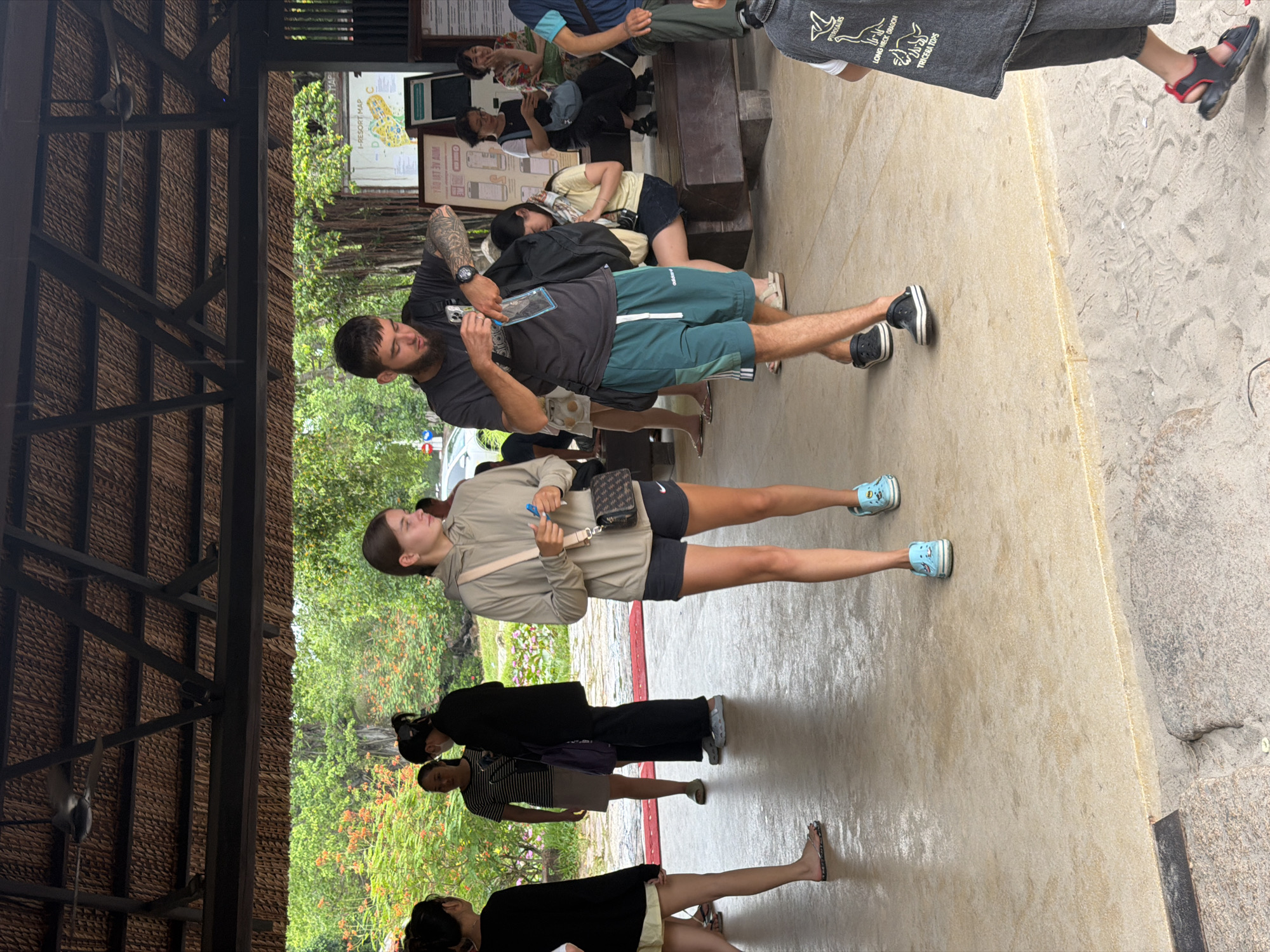 This image shows a group of people standing and walking in a covered outdoor area with a thatched roof. In the background, there are trees and foliage, suggesting a tropical or subtropical setting. A map or sign with text is visible on a bulletin board in the background. Some of the people are wearing casual clothing, like t-shirts and shorts, while others are in more subdued attire. One person in the foreground is wearing a black shirt and green shorts, and another woman is wearing a grey hooded jacket and black shorts. A few people are looking at their phones. The lighting suggests it is daytime, and the ground appears to be wet, possibly from recent rain. The scene seems to be a public area, perhaps a park, tourist attraction, or resort. The overall atmosphere appears relaxed and casual. Visible text includes "I-RESORT MAP C" and what looks like Vietnamese characters on the sign.