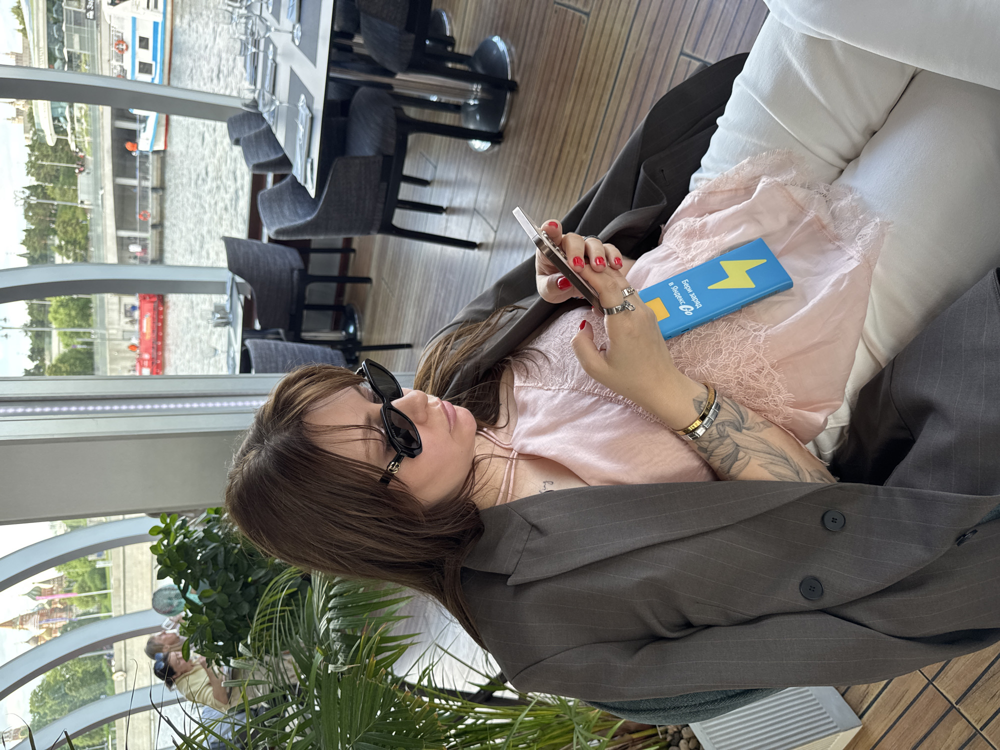The image shows a woman sitting in a restaurant, looking at her phone. She is wearing sunglasses, a blazer, and a pink top. She is also holding a blue box with a yellow lightning bolt on it. The restaurant appears to be located on a boat or near water, as evidenced by the views of the Moskva river through the windows. The buildings reflected are St. Basil's Cathedral and the Kremlin in Moscow, Russia. Several dining tables are visible, and another person is behind the plant.