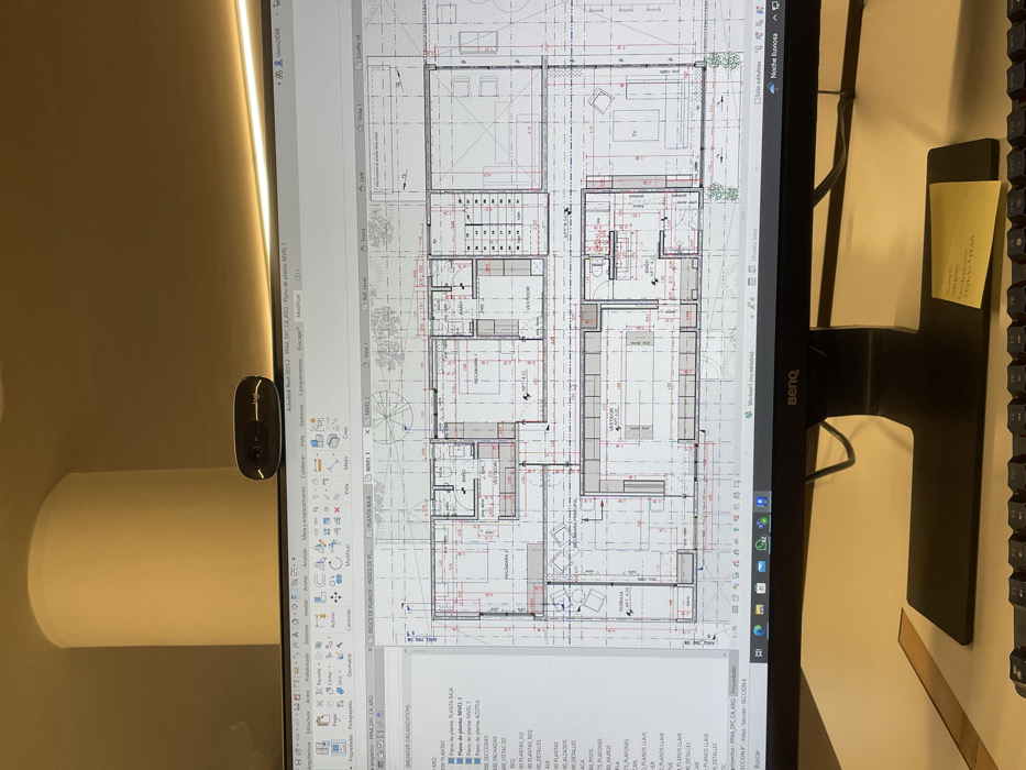 A BenQ computer monitor displays an architectural floor plan within Autodesk Revit 2023.2, showing labels such as "RECAMARA 1," "BAÑO," and "VESTIDOR," indicating a residential design. The software interface and content are in Spanish. A black web camera is mounted on top of the monitor. Below the screen, a partially visible black keyboard and a yellow sticky note with handwritten text ("SOLICITUD:") are present on a desk. Overhead, a cylindrical, off-white light fixture is visible. The scene depicts an indoor architectural workspace, located in Mexico City, Mexico.