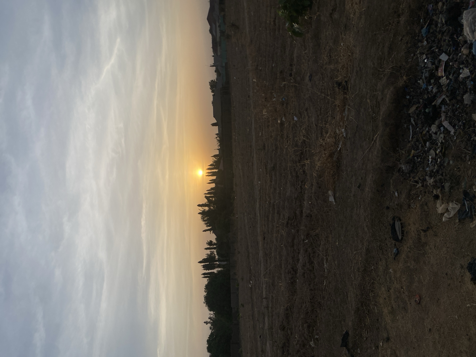 The image depicts a sunset over a field in Rido, Nigeria. The sun is setting on the horizon, casting an orange glow on the sky. There are some trees and bushes in the background. The field is covered in dry grass and some trash. The sky is overcast with light clouds. There are no people visible in the image.
