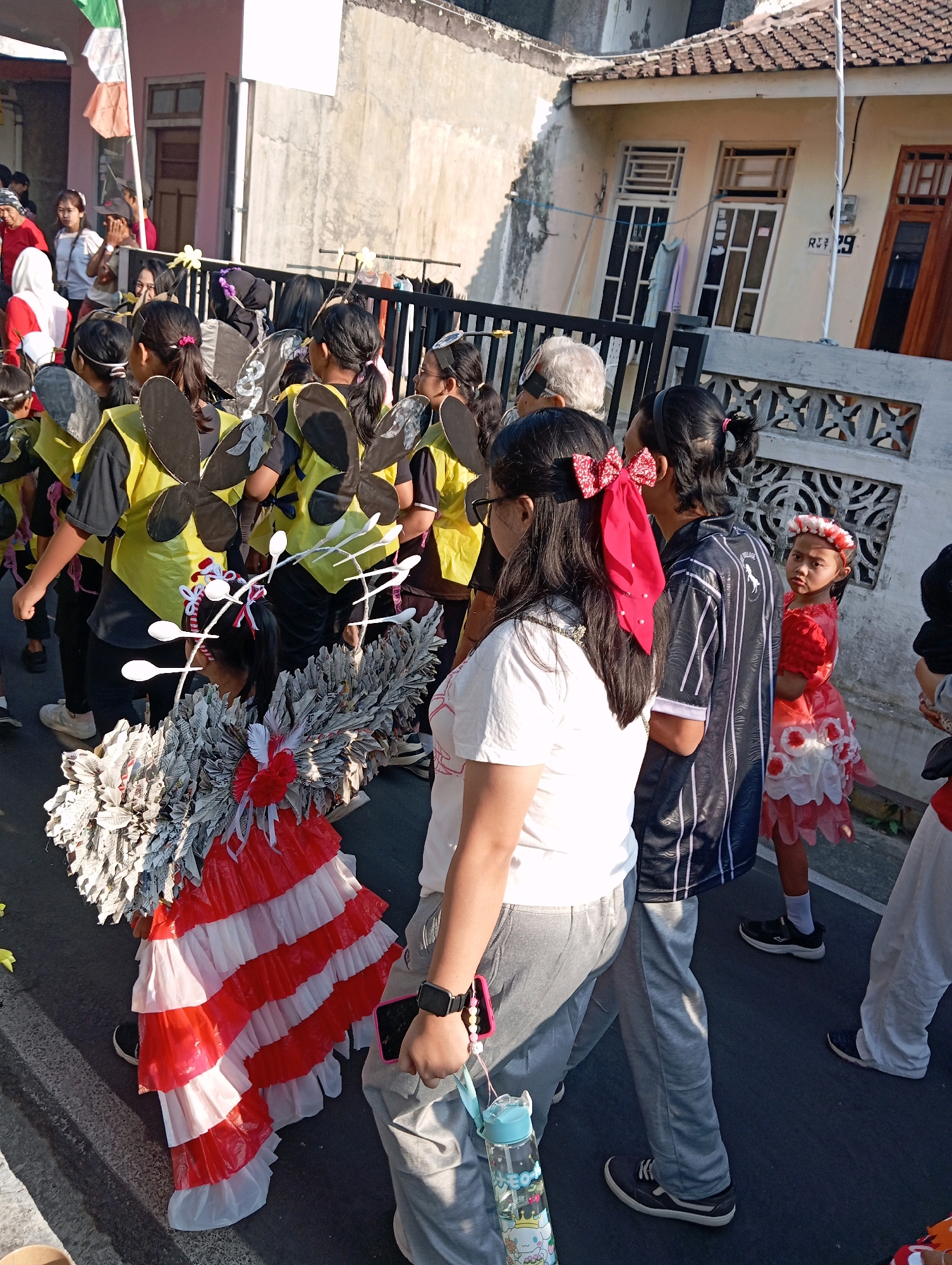 In Magelang, Indonesia, a group of children dressed in bee costumes participates in a parade. The children are wearing yellow and black outfits with large, black, butterfly-like wings. One child in the foreground wears an elaborate costume resembling a bee with red and white ruffled layers and large, textured wings. Other participants are visible in the background, including an adult with white hair and a woman in a black and white striped shirt. A young girl in a red dress and floral headband stands to the right, looking towards the camera. The parade takes place on a paved street, with buildings and fences lining the background. The lighting suggests it is daytime, possibly late afternoon, casting shadows on the street.