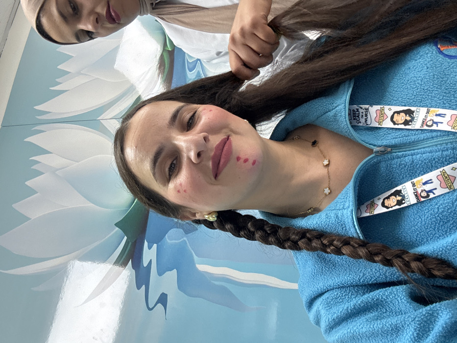 This scene occurs indoors in Meknes, Morocco, depicting two women. The primary subject, positioned in the foreground, is a woman with long, dark brown hair. Her left side features a completed braid, while her right remains unbraided. She is observed wearing a light blue zippered fleece jacket, a gold necklace adorned with white clover motifs, and gold hoop earrings. Red marks are present on her upper cheeks and chin. A white lanyard displaying text such as "KEEP CALM I'M A NURSE" and "DANCE IT OUT" is suspended from her neck. A second woman, partially visible in the background and wearing a light-colored hijab and a white garment, grasps a section of the primary subject's unbraided hair, indicating an ongoing or impending hair braiding activity. The background consists of a wall adorned with a mural depicting stylized white lotus flowers floating on a light blue surface with undulating water patterns.