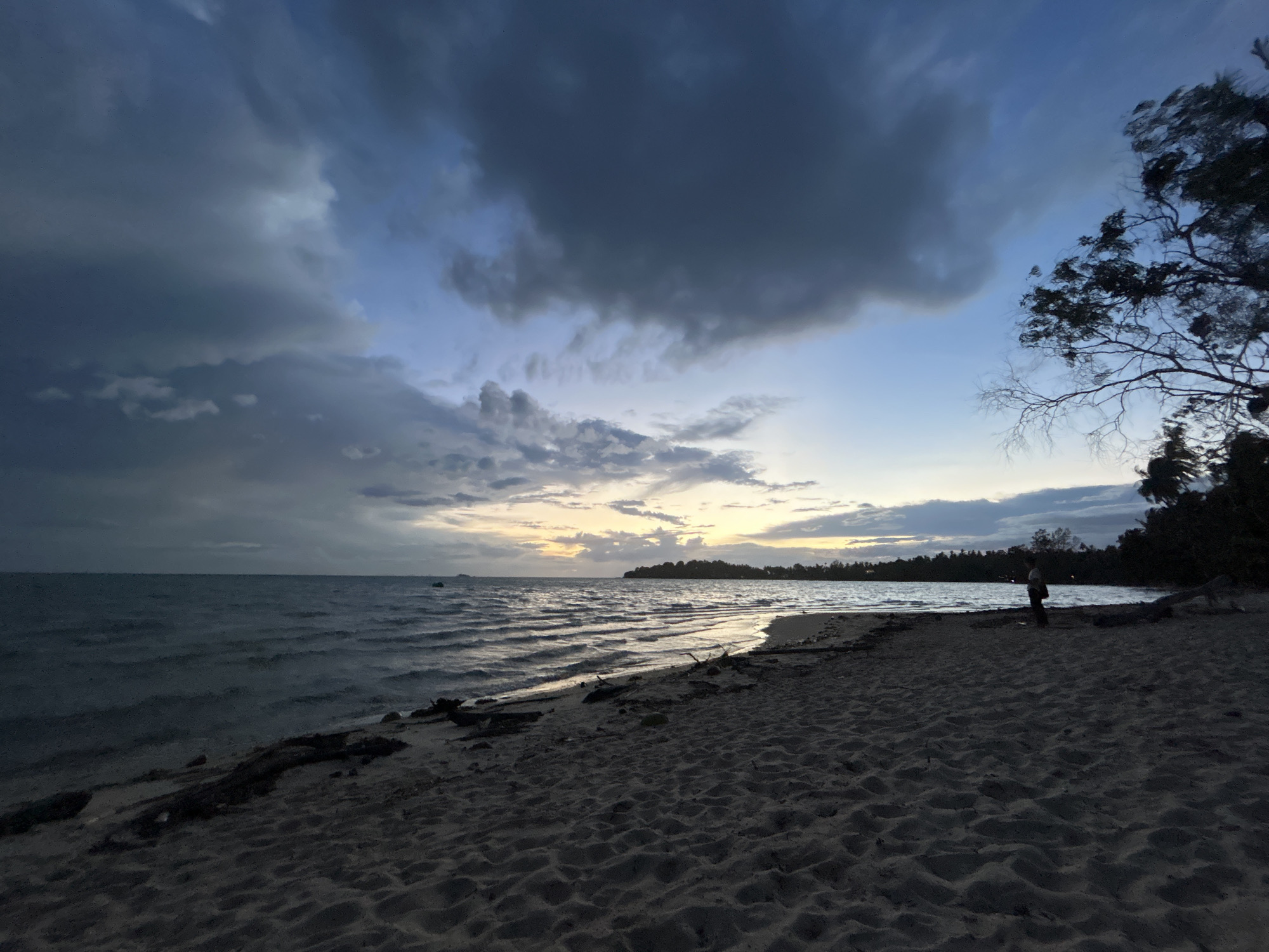 This is a landscape photograph taken at dusk on a sandy beach in Koh Phangan, Thailand. The scene is dominated by a dramatic sky filled with dark, textured clouds, interspersed with patches of lighter blue and a faint glow of sunset on the horizon. The sea is choppy, with small waves rolling onto the shore, reflecting the muted light of the sky.
On the right side of the frame, a lone figure stands silhouetted against the sea, facing towards the water. The beach is covered in sand and scattered with driftwood. The overall atmosphere is one of a coming storm or the lingering light after one has passed, with a sense of quiet contemplation. The time of day is clearly evening, transitioning into night. The location is identified as Koh Phangan, Thailand. There is no visible text in the image.