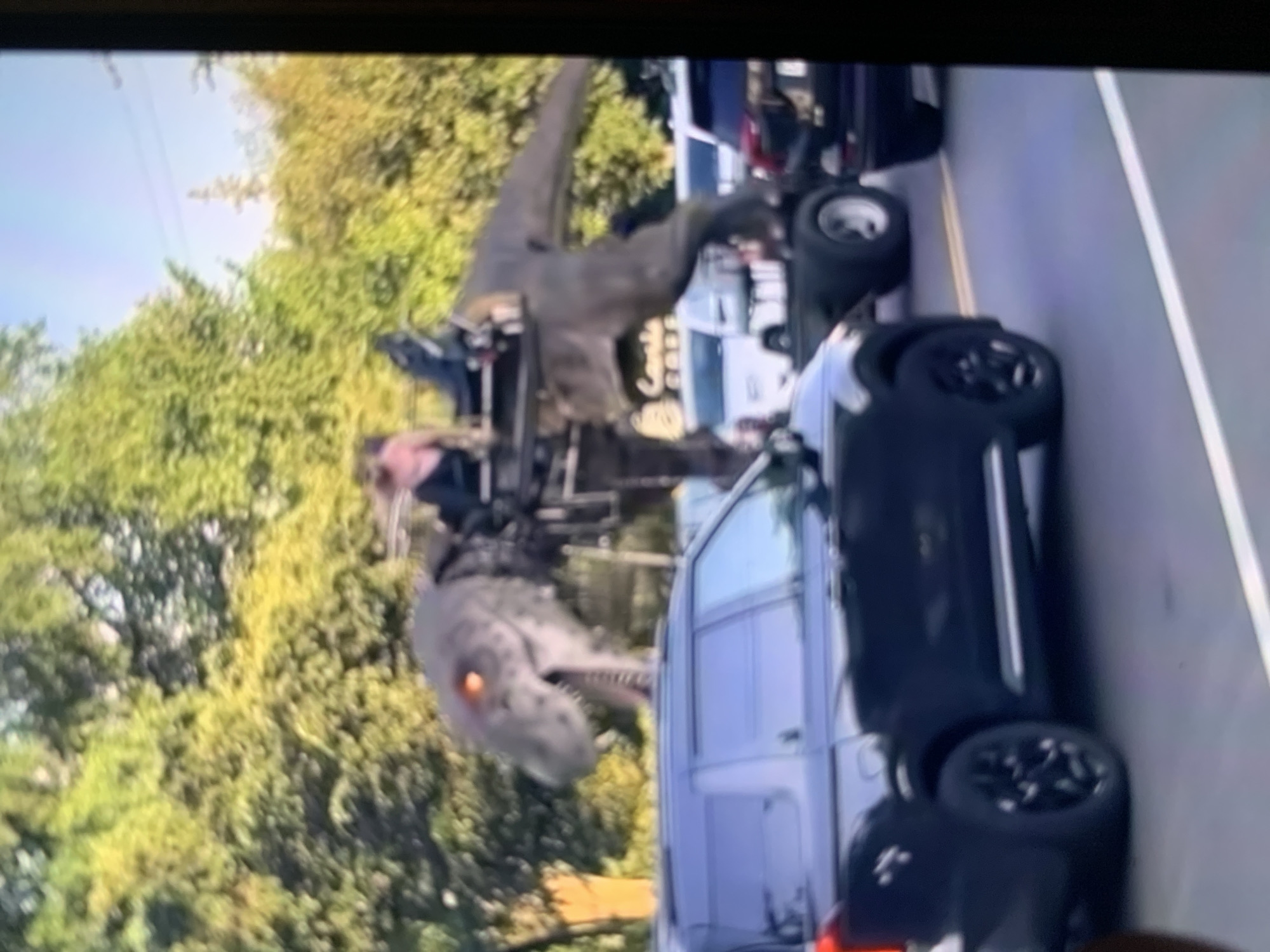 The image depicts a scene that appears to be from a movie or television show, featuring a large, realistic dinosaur prop in an outdoor setting. The dinosaur is mostly gray with a textured skin and has a menacing appearance, with glowing orange eyes. It is positioned on a trailer or platform that is being towed, suggesting it is part of a performance or display.
In the foreground, a black SUV is visible, with its wheels and side clearly in view. The SUV is parked on a road. To the left of the dinosaur, a silver van is also present. There are people in the scene, some of whom are near the dinosaur on the trailer, appearing to control or manage it. The overall setting suggests a suburban or semi-rural environment with trees and foliage in the background.
The image is somewhat blurry and low-resolution, making it difficult to ascertain specific details about the time of day or weather conditions, though the lighting suggests it could be daytime. There are no clear location cues that specifically point to Stavropol, Russia, as the image itself does not contain any identifiable Russian landmarks or text. The presence of the dinosaur prop suggests an event that is likely for entertainment purposes, such as a parade, festival, or filming.