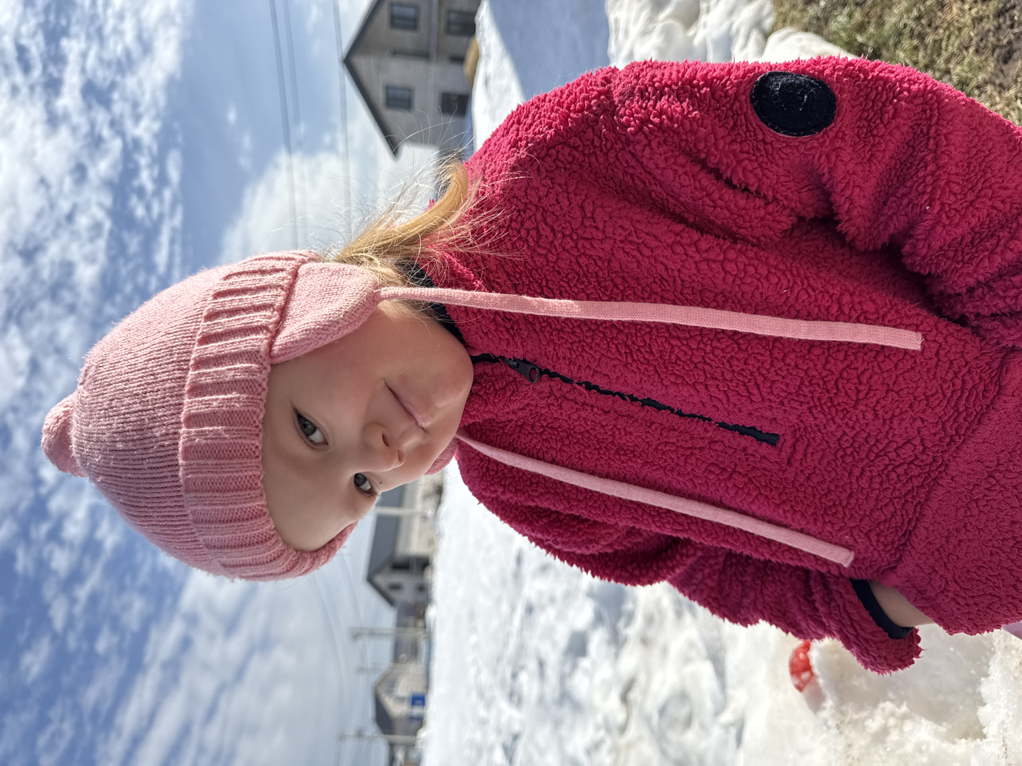A young child is depicted outdoors in Gornyi (Dvurechenskoe S/P), Russia. The child, situated in the foreground, wears a light pink knitted winter hat with ear flaps and ties, and a fuchsia-colored textured jacket with a black half-zip closure. A circular dark patch is visible on the jacket's right sleeve. The child looks directly forward with a slight, closed-mouth smile and a tilted head. The scene is illuminated by direct sunlight under a blue sky with scattered white clouds. The background features a residential landscape covered in melting snow, revealing patches of brown grass. Multiple two-story houses with light-colored facades and dark roofs are visible, along with utility poles and lines. A small red object is partially obscured in the snow to the child's lower left.