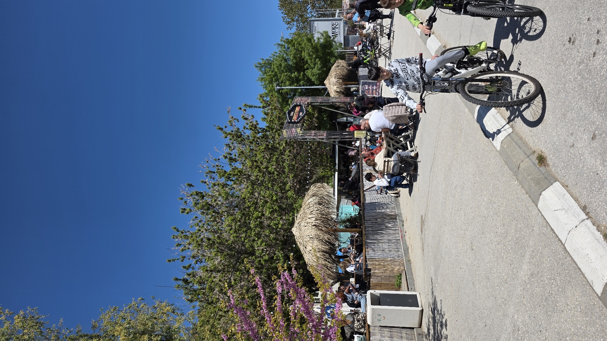 The image captures a bustling scene along a street in Varna, Bulgaria, characterized by the presence of outdoor dining establishments and pedestrian activity. A group of people is seated at tables under a thatched-roof structure that appears to be part of an outdoor café or restaurant. Signage is visible for a place called "Street Food Bar". Patrons are seated on folding chairs and appear to be engaged in conversation and dining. Cyclists are seen riding on a road to the right.
Trees and flowering vegetation line the left edge of the street, adding a touch of greenery to the urban setting. The sky above is clear and blue. The overall impression is a vibrant outdoor environment.