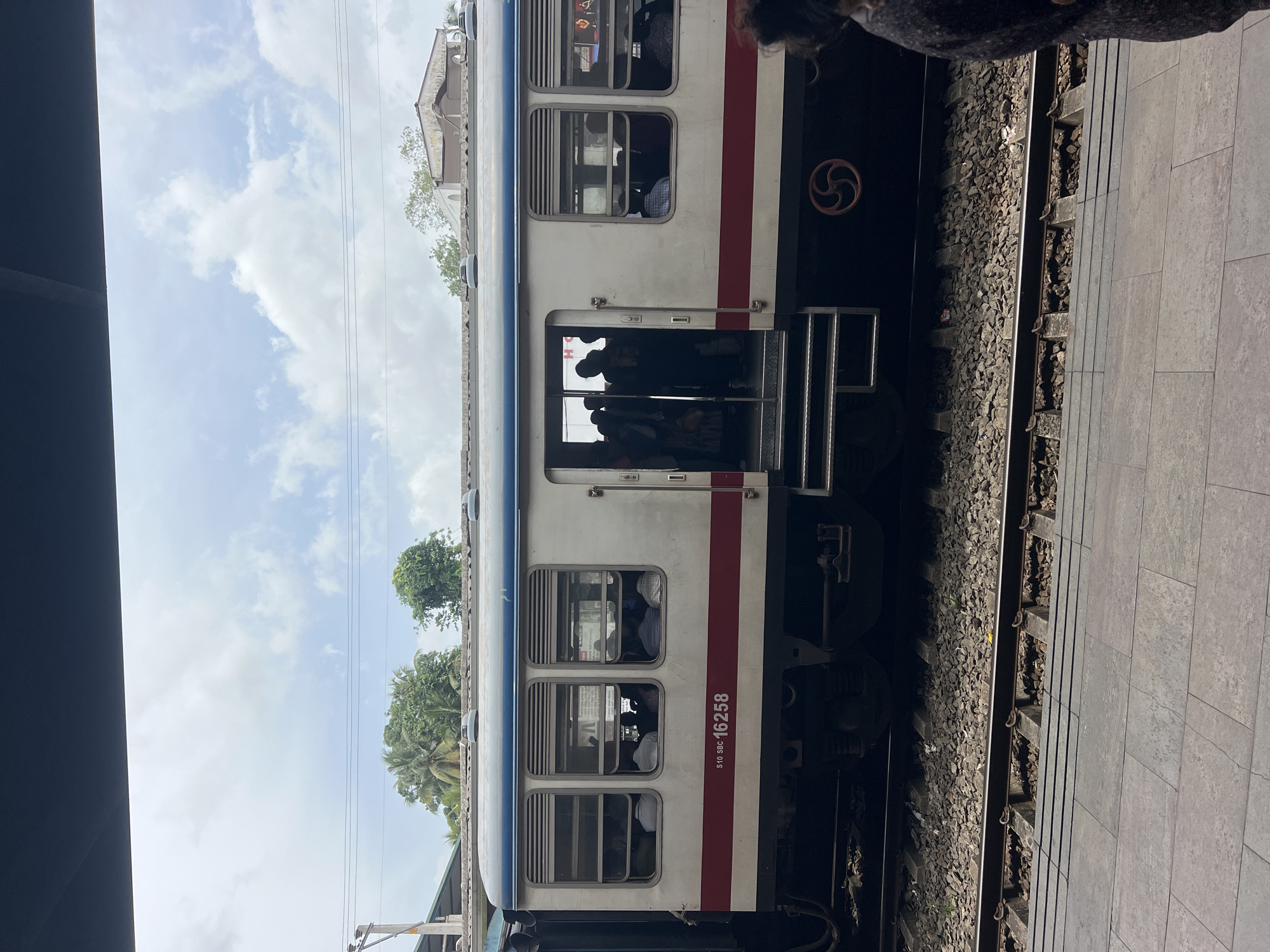 The image shows the side of a train carriage at a station, likely in Gampaha, Sri Lanka, due to the presence of palm trees and the architectural style of the buildings in the background. The train is white with a blue and red stripe along its side. The number "510 SBC 16258" is visible on the carriage. People are seen inside the train, and some are standing in the open doorway, suggesting it is either boarding or disembarking. The train is on a set of tracks, with ballast visible between the sleepers. A platform, paved with large, square tiles, is in the foreground. The sky is partly cloudy, and the overall lighting suggests daytime.
