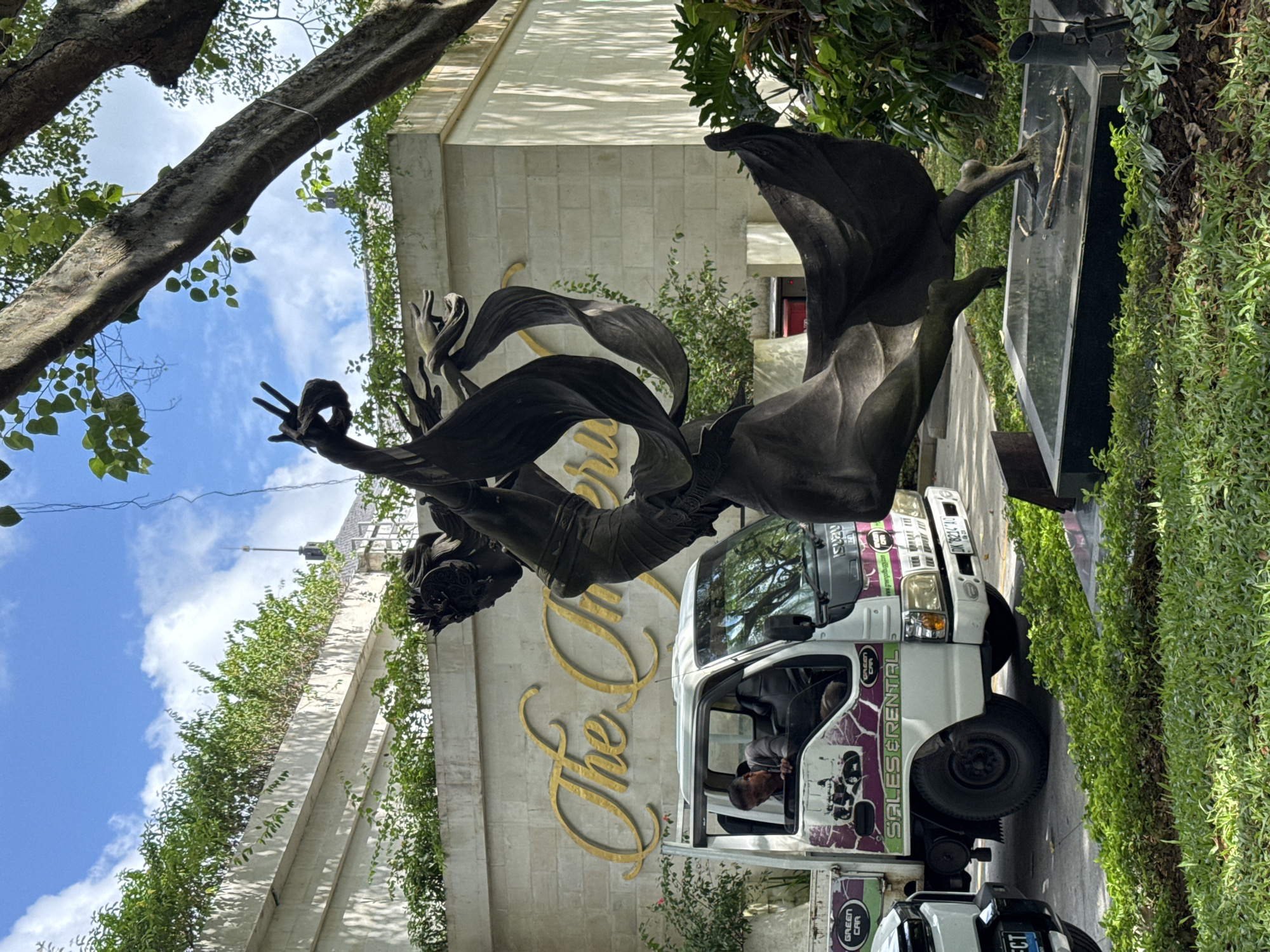A medium-angle, candid outdoor shot shows a statue of a female figure with flowing fabric, a "The Ritz-Carlton" sign on a cream-colored wall, and a white truck with the words "SALES RENTAL" and "GREEN CAR" on the side, taken in Kuta Selatan, Indonesia.
The statue is positioned prominently in the foreground. The figure is dark bronze, possibly metal, and appears to be in mid-motion with an arm raised and a gesture. It is set against the backdrop of "The Ritz-Carlton" sign, written in elegant golden script.
A white truck is parked adjacent to the statue. The driver, a dark-haired man, is visible in the driver's seat. The truck has distinctive graphics and lettering, with the phrases "SALES RENTAL" and "GREEN CAR" clearly visible.
The building behind the statue is cream-colored and has an elaborate design, with some portions covered in greenery. Trees with leaves visible are present around the building and the statue, and the sky is visible through the tree branches.