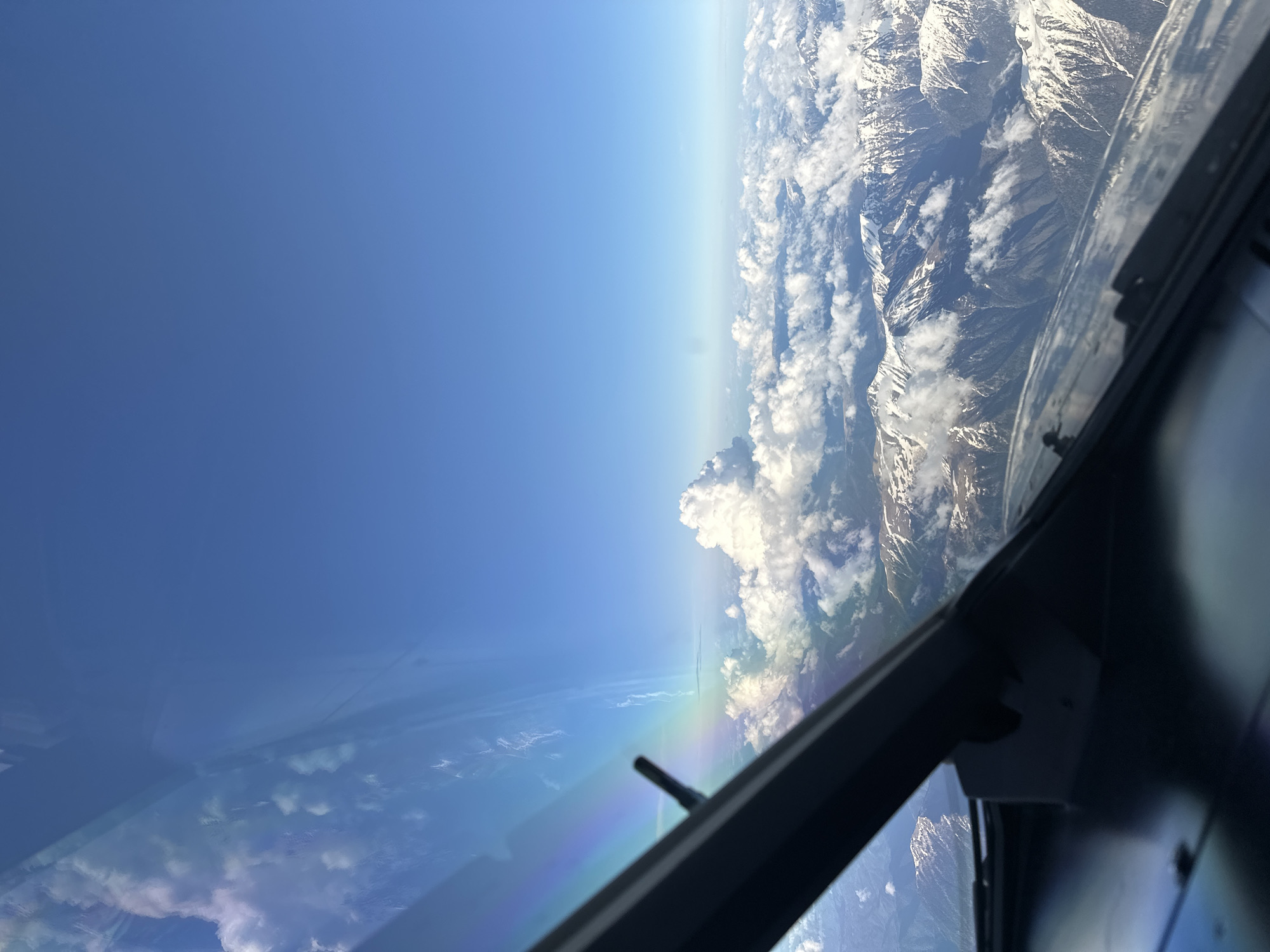 The image shows a view from inside the cockpit of an aircraft. The aircraft is flying above a landscape consisting of mountains partially covered with snow and clouds. Outside the window, there is a bright blue sky with an atmospheric haze. A rainbow effect is visible on the cockpit window. The location is not discernible from the image. There is no evidence to suggest that the image was taken near the city of Khimki, Russia.