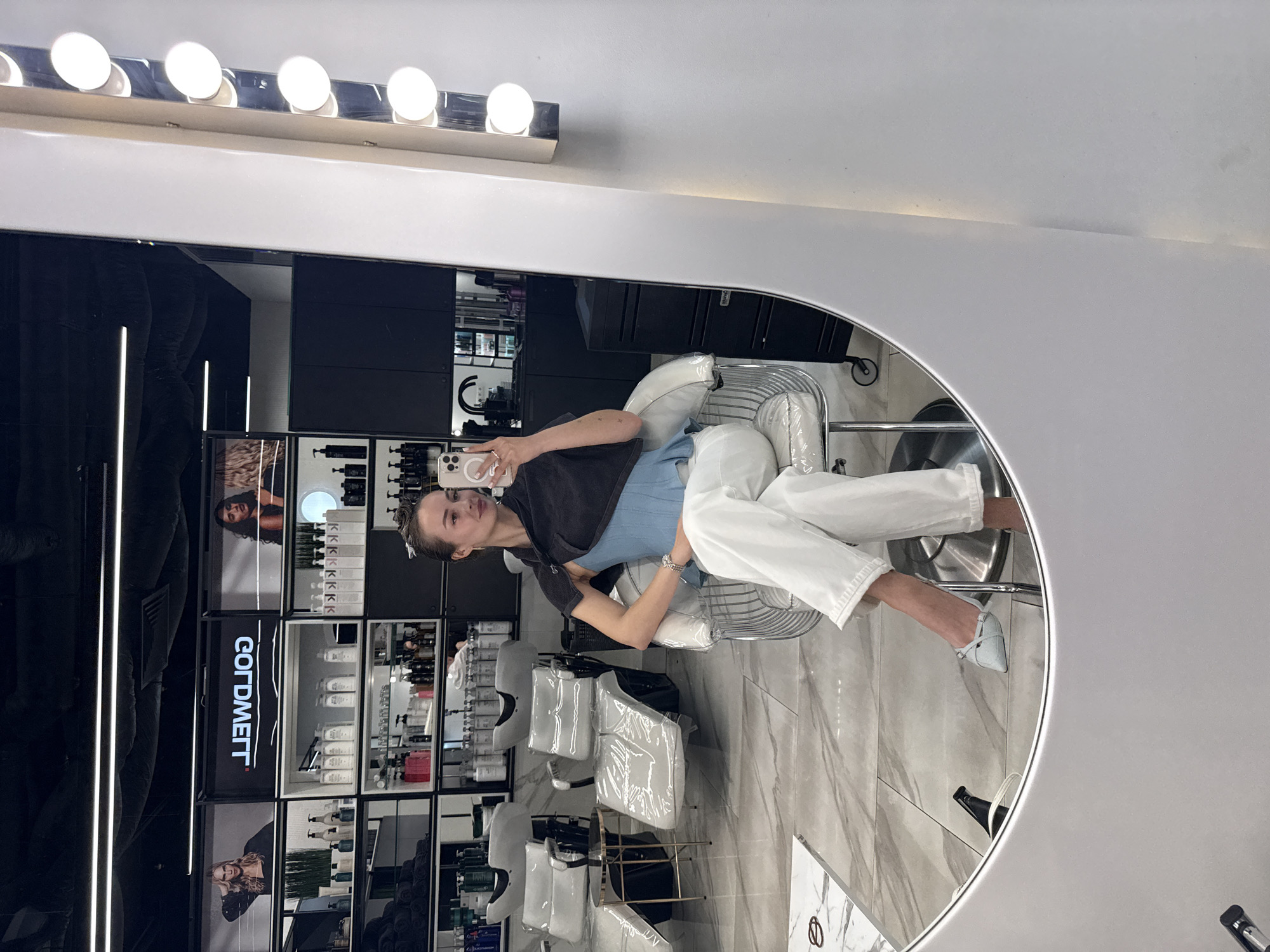 A woman is reflected in a circular mirror in a salon, holding a cell phone to take a picture. The salon has a shelf with "GOLDWELL" printed on it. Other chairs are present. The setting is likely a hair salon in Kyiv, Ukraine, based on brand and cultural context.