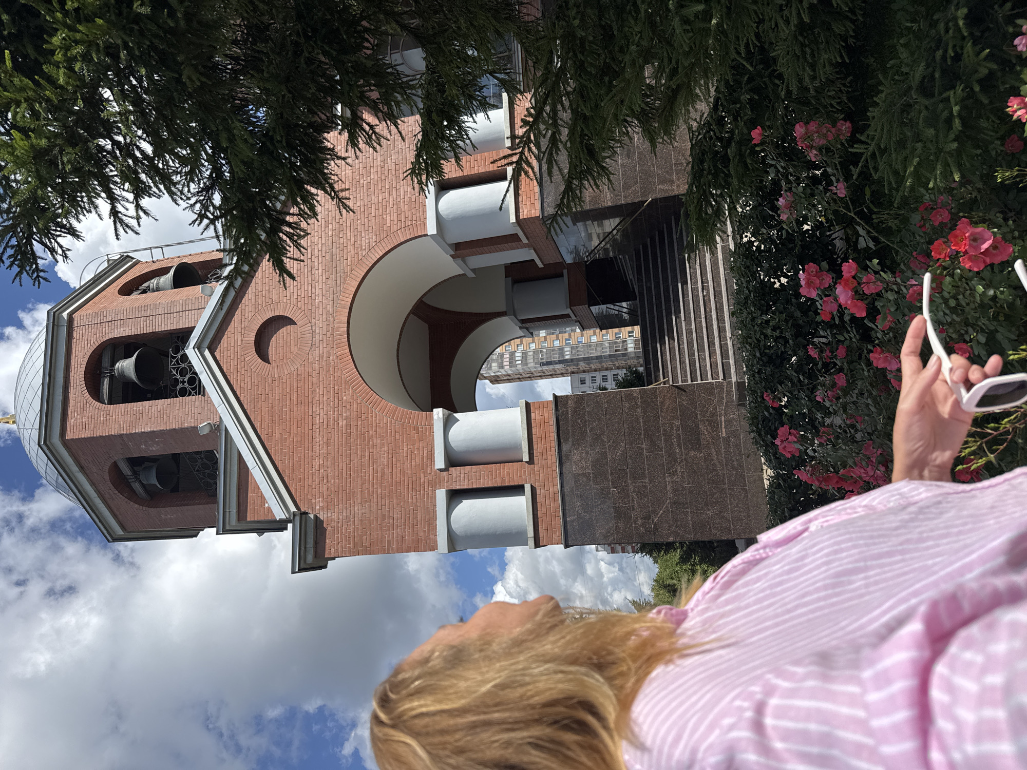 A woman with blonde hair, wearing a pink and white striped shirt, is viewed from behind and to the side. She is looking upwards towards a bell tower. The bell tower is constructed of red brick with arched openings. Inside the openings, bells are visible. There is a dome on top of the bell tower. In front of the tower, there are stairs leading up to an arched entryway. A garden with pink roses is situated in the foreground. A large evergreen tree is to the right of the frame, partially obscuring the bell tower. In the background, a modern apartment building can be seen. The sky is blue with white clouds. The scene appears to be a sunny day.