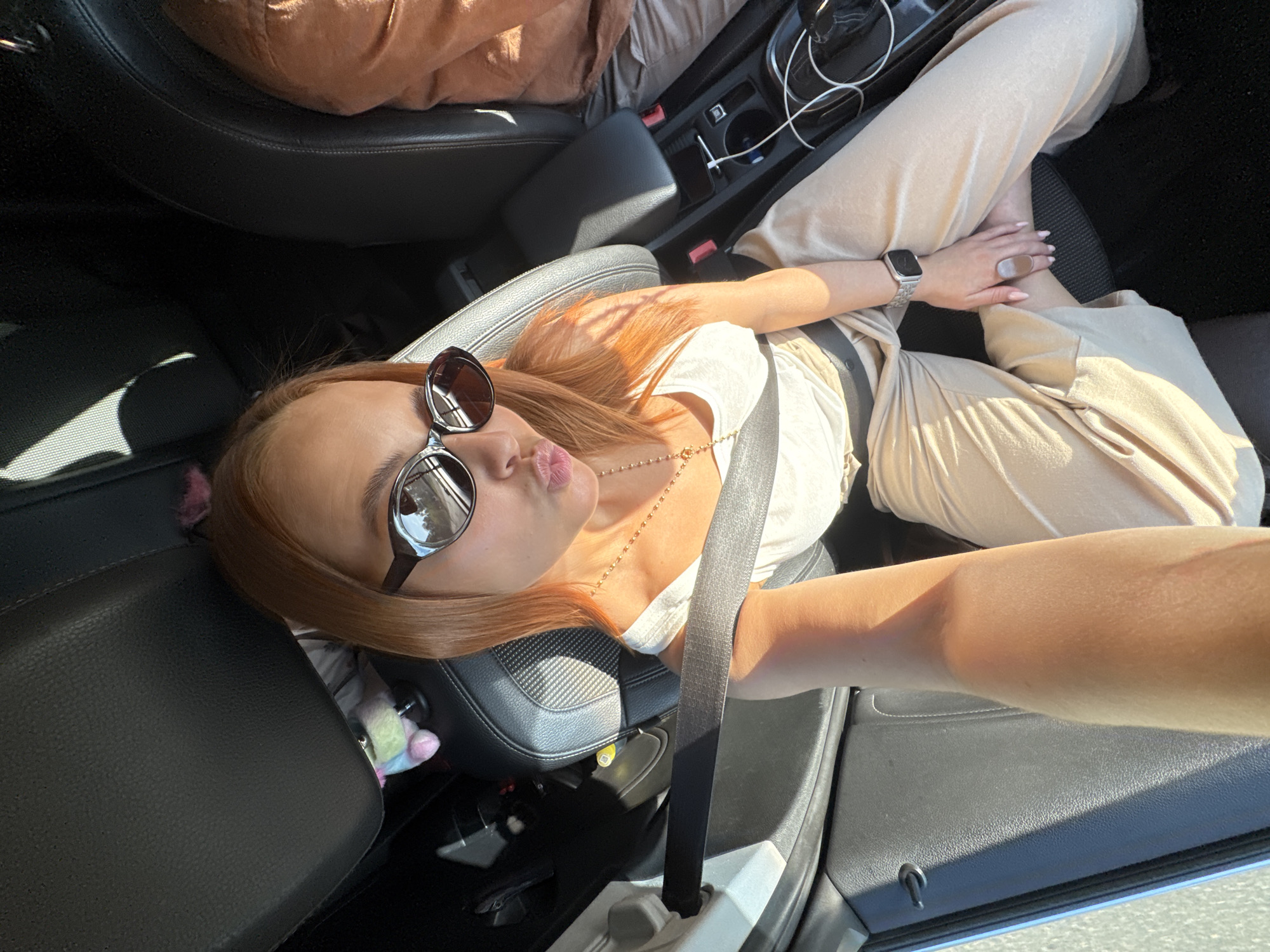 A woman with auburn hair, wearing a white tank top, beige pants, sunglasses, and a silver watch, is seated in the driver's seat of a car. She is holding her phone with her right arm to take a selfie, puckering her lips. A seatbelt is fastened across her chest. The interior of the car is visible, including the black leather seats and the center console with a charging cable. Sunlight streams into the car, casting shadows on the interior. The location is identified as being in the city of Chaikovsky, Russia.