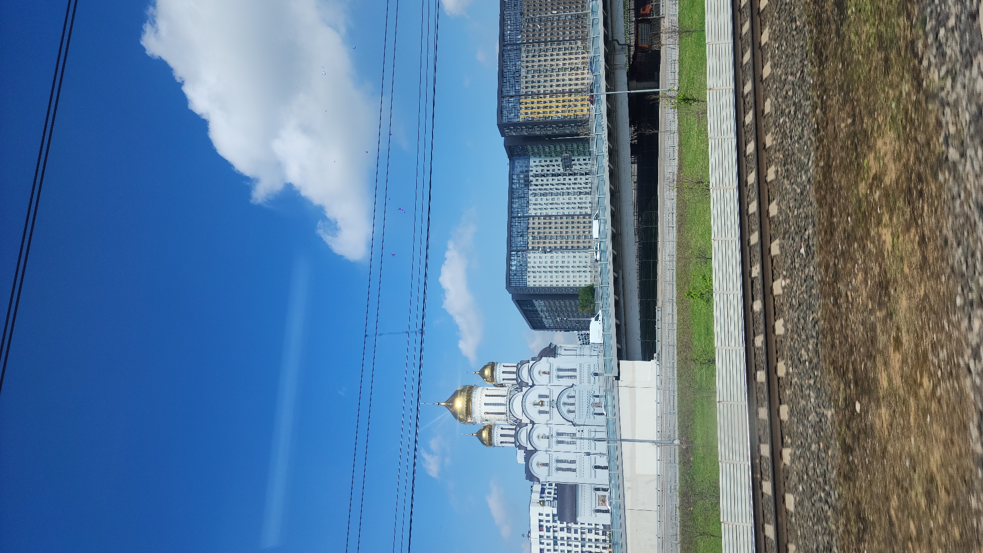 The image depicts a cityscape under a clear blue sky with scattered clouds. Dominating the view is a large white building with multiple golden domes, likely an orthodox church. Adjacent to the church are modern high-rise apartment buildings. A set of train tracks runs alongside the buildings, separated by a narrow strip of grass. Overhead power lines stretch across the sky. The image appears to have been taken from a moving train. The location is not specified, making it impossible to determine the city.