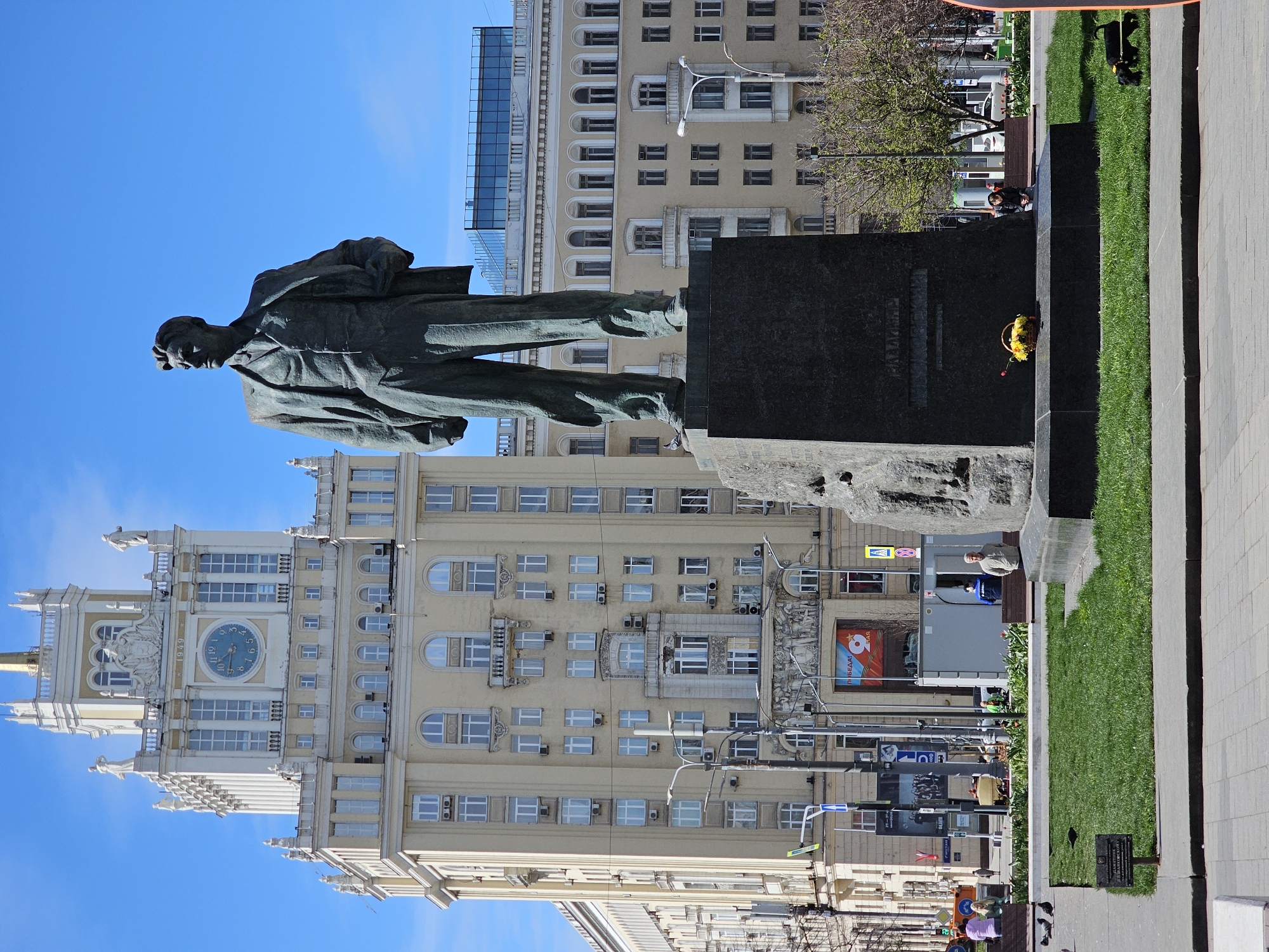 The image shows a statue of Yuri Andropov in Moscow, Russia. The statue stands on a plinth in front of a building with a clock tower. There is a small park area near the statue, and several people are walking in the background.