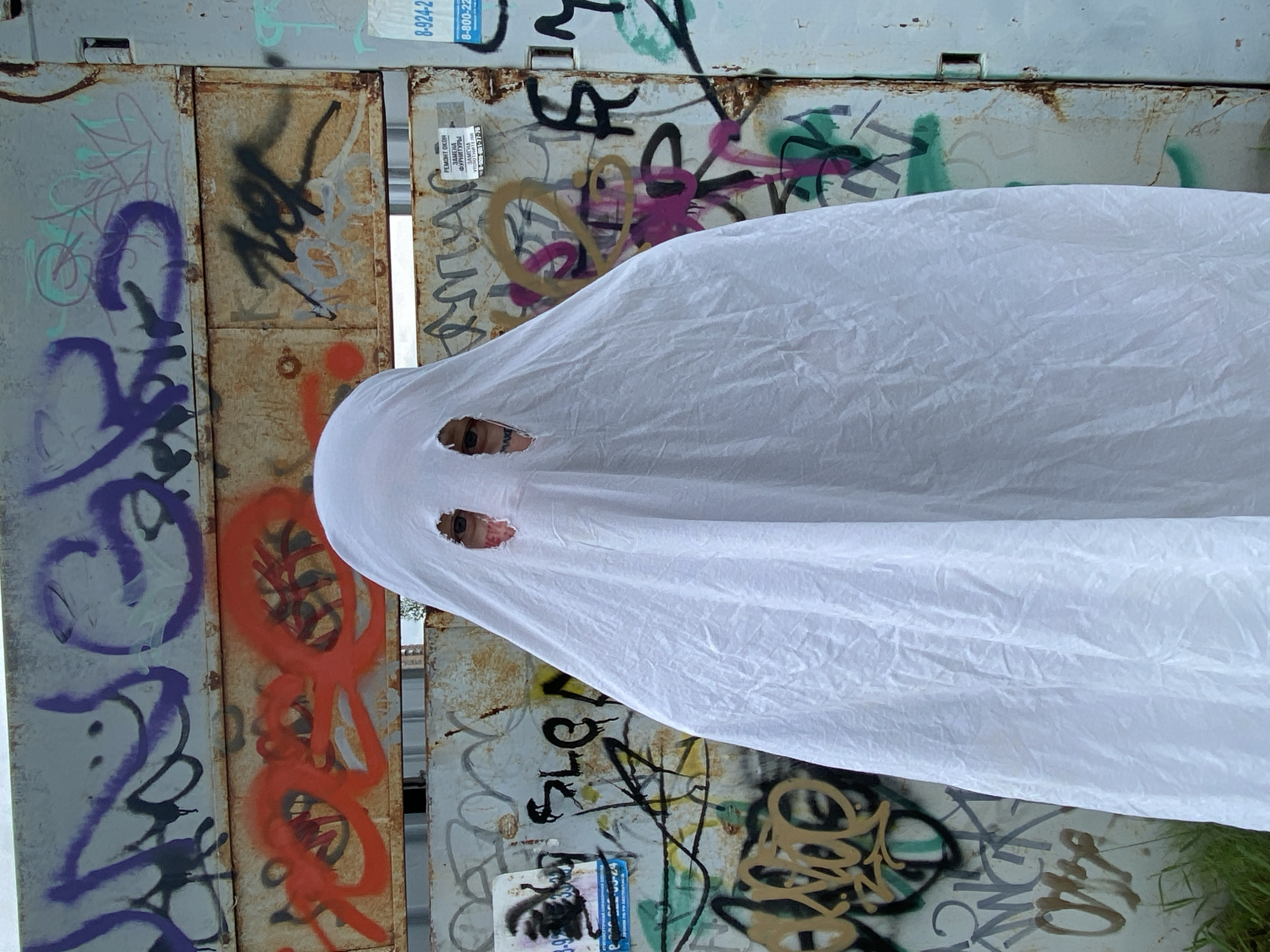 A person dressed as a ghost stands in front of a graffiti-covered metal wall in Khabarovsk, Russia. The ghost costume is a simple white sheet with two eyeholes cut out, revealing the person's eyes. The wall behind the ghost is covered in various tags and graffiti in different colors, including purple, orange, black, and green. A few flyers are also stuck to the wall. The ground in front of the wall is covered in grass and dirt. The scene appears to be outdoors, possibly in an urban alleyway or industrial area.