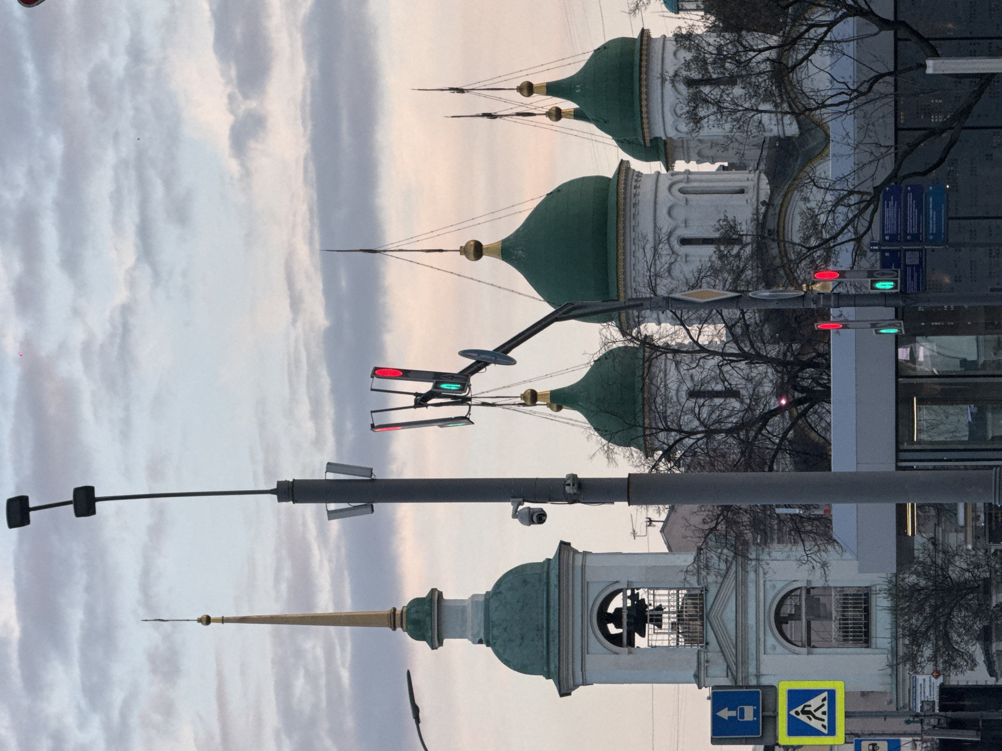 The image shows a view of a street in Moscow, Russia. In the foreground, there are several streetlights and traffic signals. The traffic lights show both red and green signals illuminated. Further back, there are architectural structures, including buildings with domes, indicative of Russian Orthodox churches or cathedrals. Bare tree branches are visible against the buildings and the sky. The sky itself is a mix of light and dark clouds, suggesting either sunrise or sunset. Street signs with directions are also visible in the lower part of the frame.